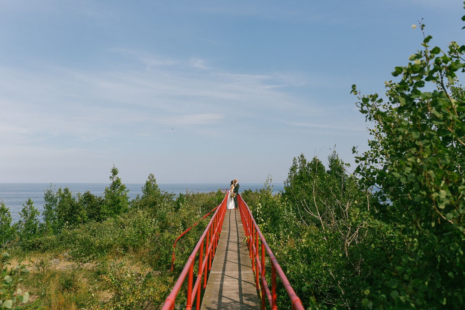 A scenic red wooden boardwalk stretches across natural grasslands under blue skies.