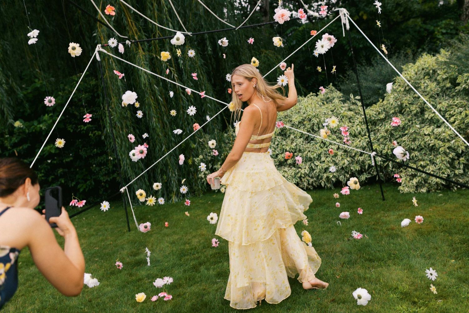 A bride in a flowing cream dress dances under strings of hanging flowers in a lush garden setting.
