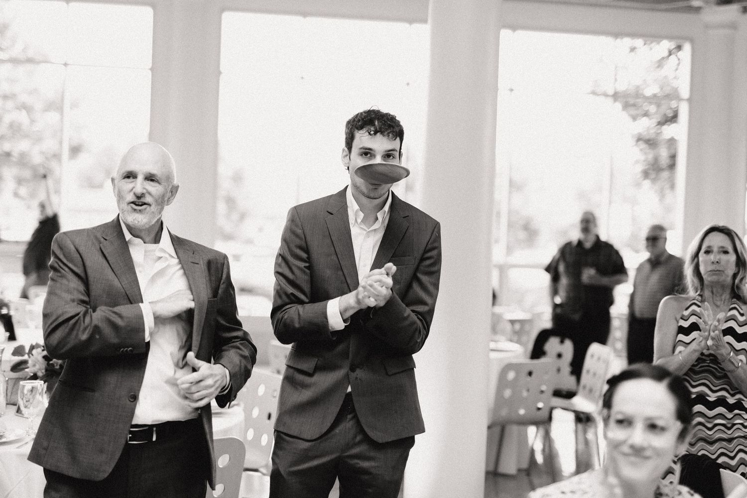 Two men in suits share a laugh during an outdoor event under white columns.