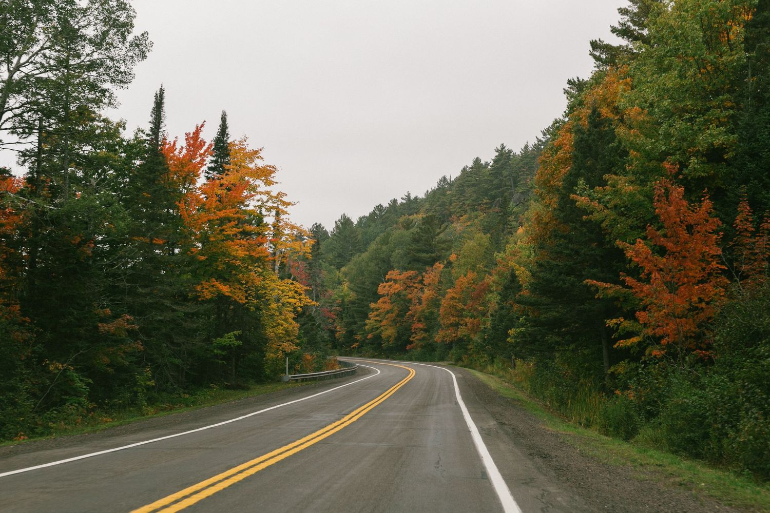 A winding rural highway curves through autumn forest with orange and red foliage in New England.