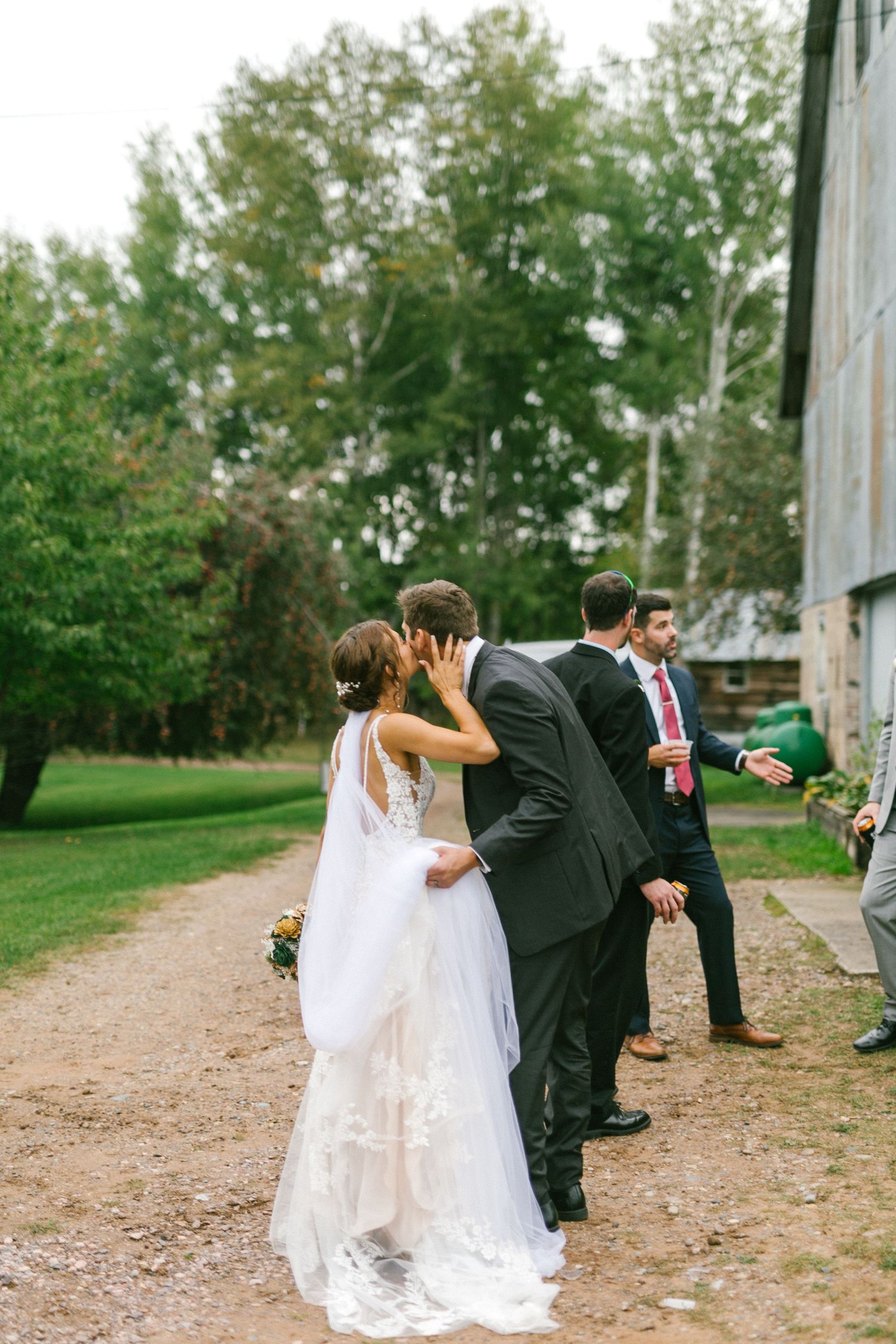 Wedding couple shares an intimate moment on a rustic dirt path near a barn surrounded by green trees.
