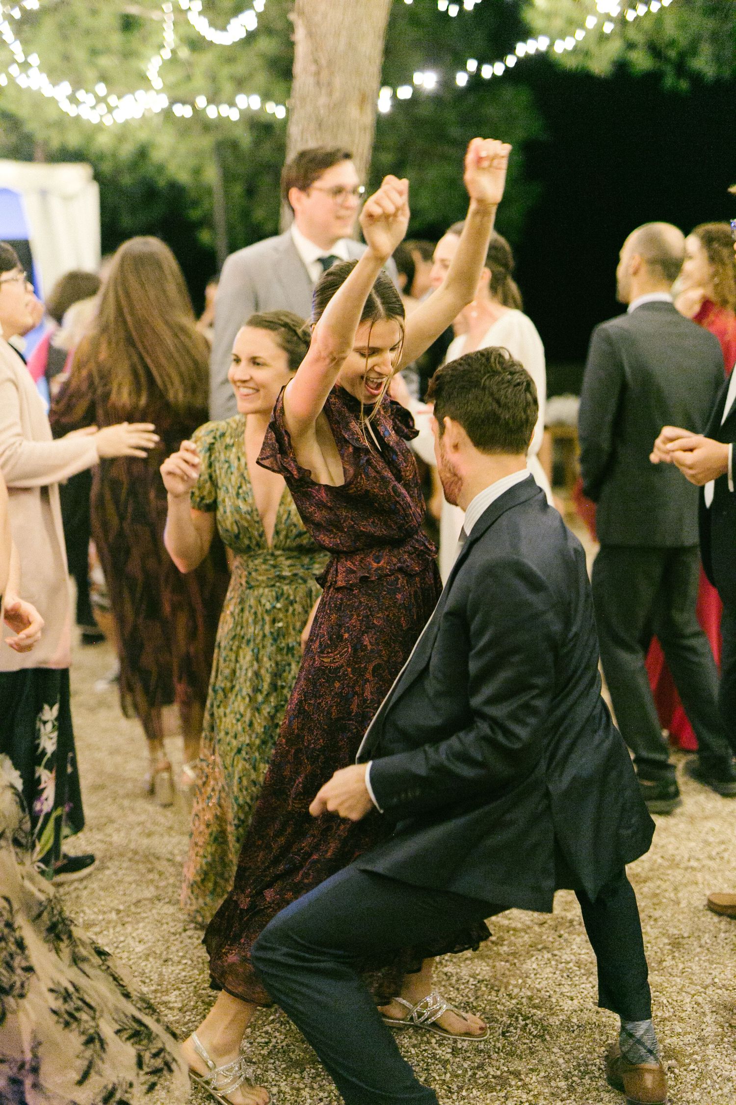 Guests dance joyfully at an outdoor wedding reception decorated with string lights at dusk.
