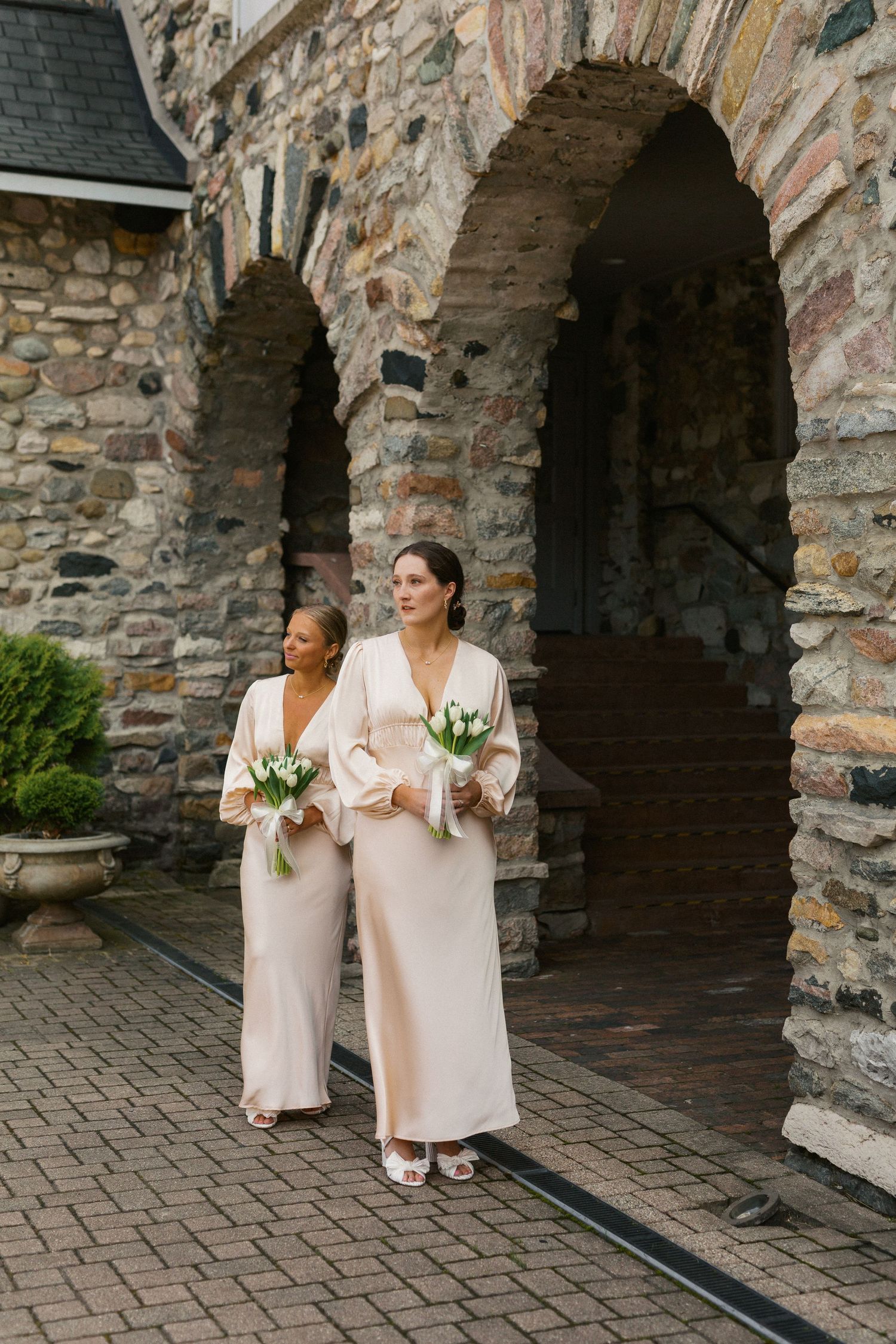 Two bridesmaids in blush pink jumpsuits holding bouquets stand against a stone archway.