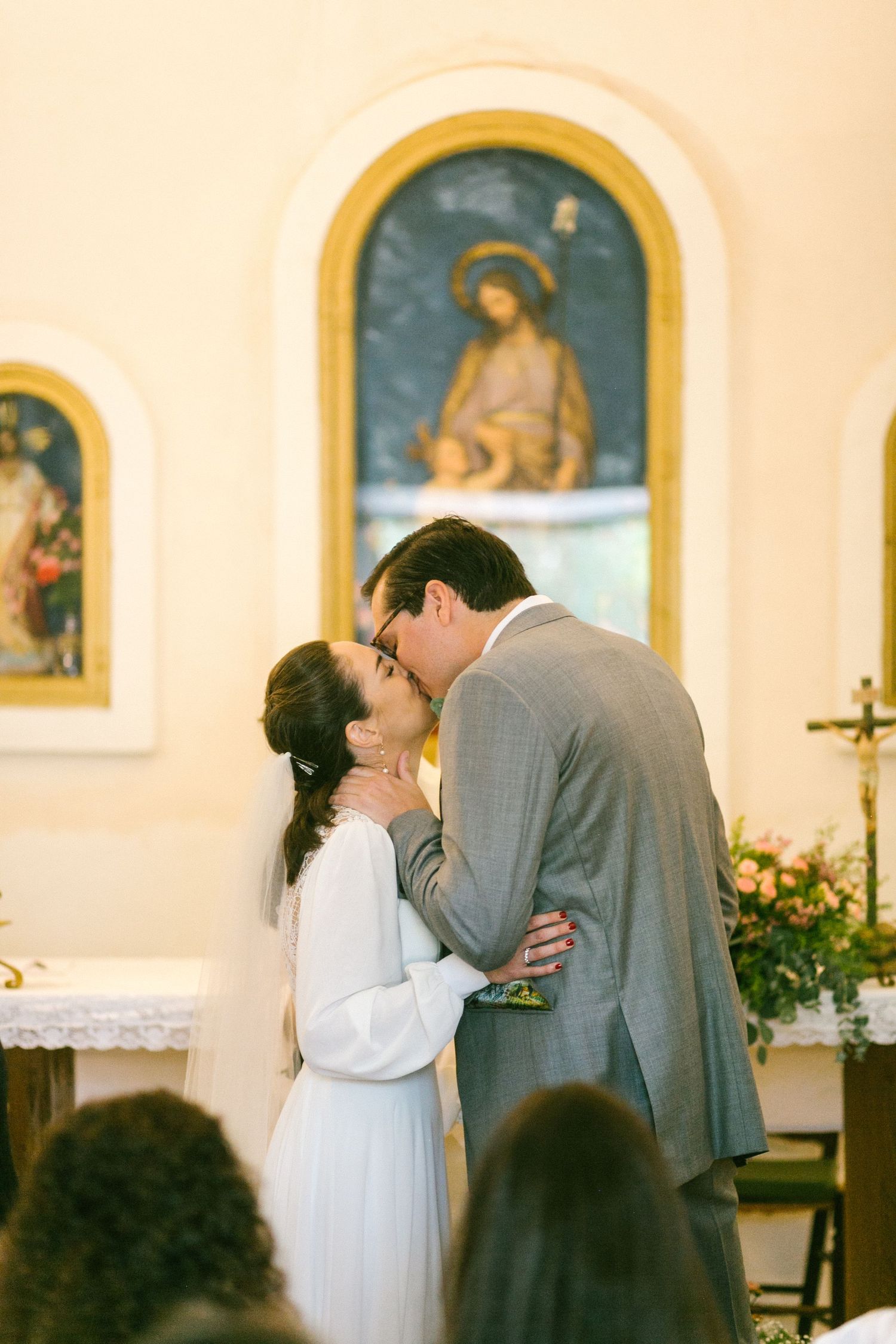 A couple shares their first kiss during a Catholic church wedding ceremony beneath religious artwork and icons.