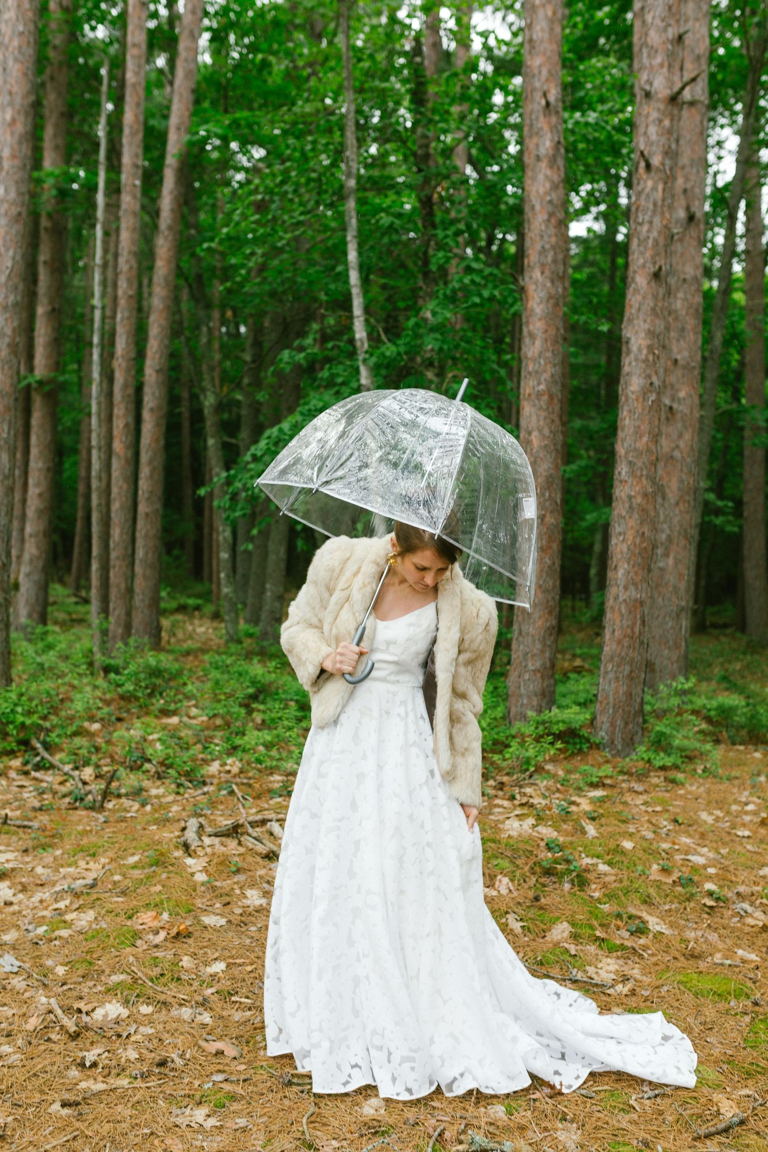 Someone in a flowing white lace wedding dress holds a clear umbrella while standing in a forest setting.