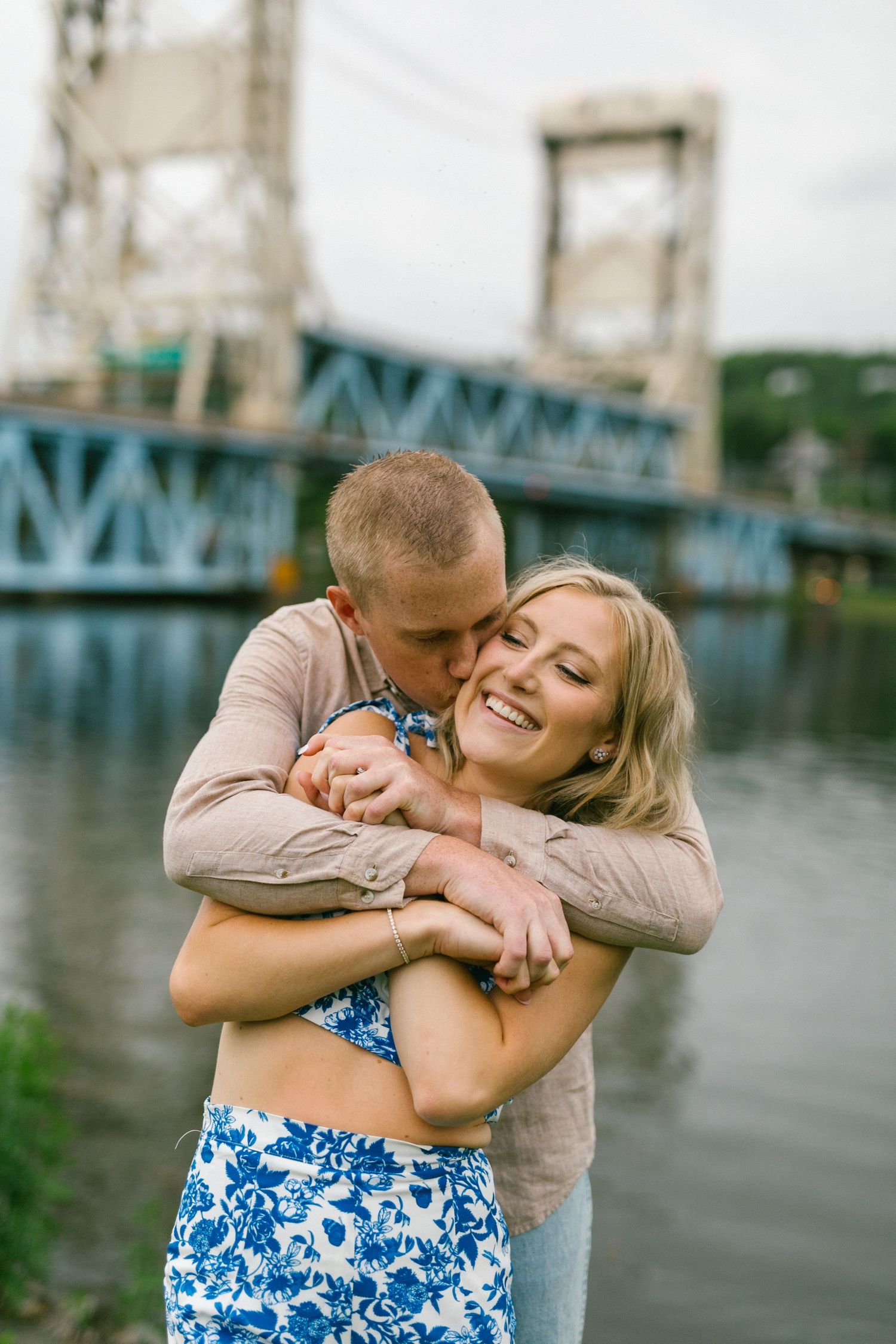 Romantic couple embraces by a blue bridge over water, with her wearing a floral blue dress and him in beige.