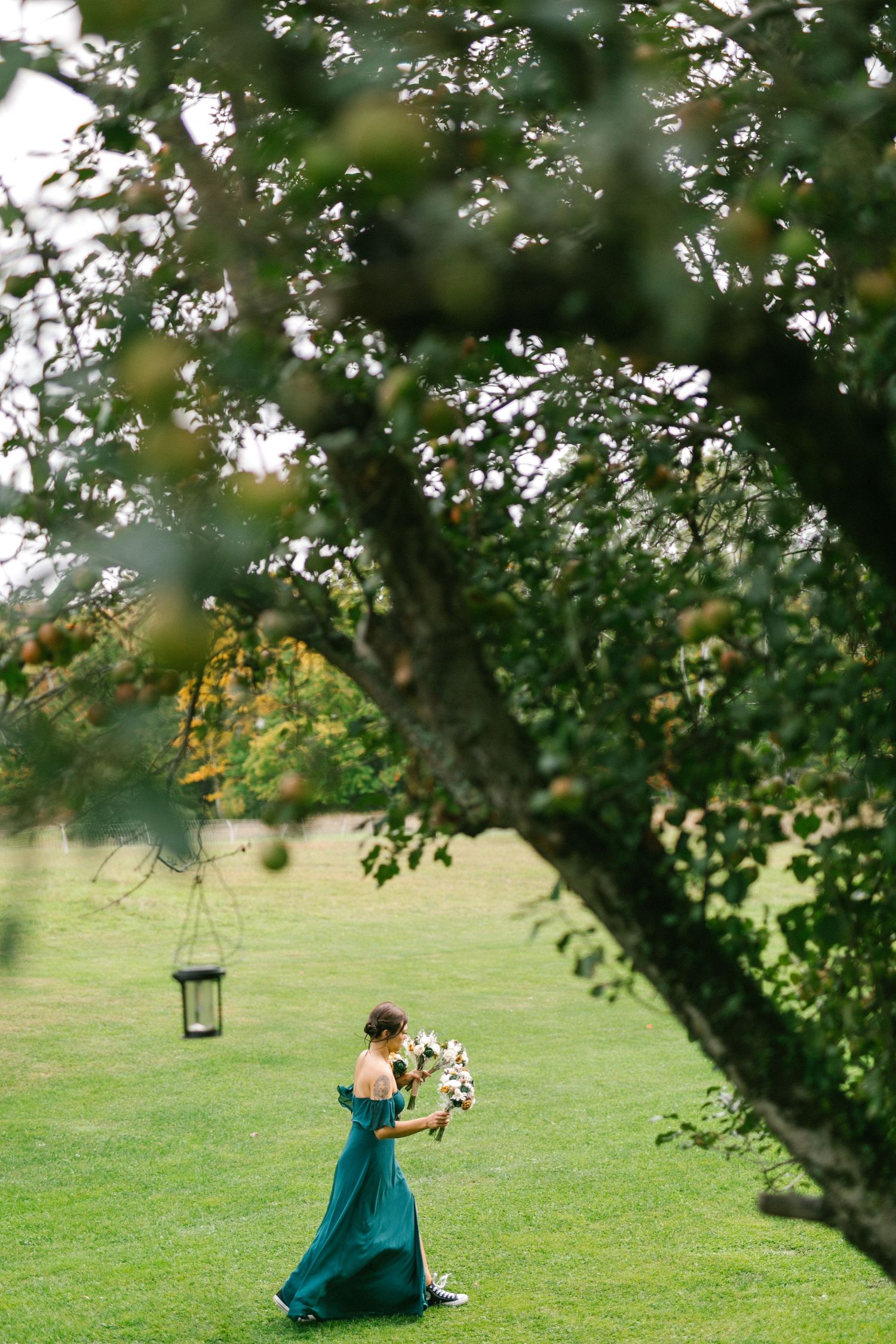 A maid of honor in a flowing teal gown holds a bouquet while walking across a green lawn under a large tree with a hanging lantern.