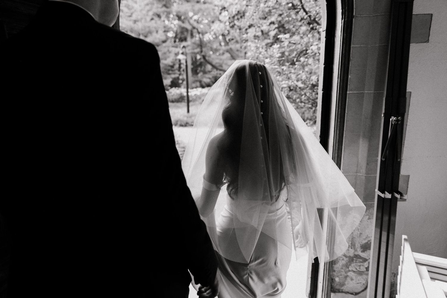 Black and white photo of a bride in veil and groom standing at an open door on their wedding day.