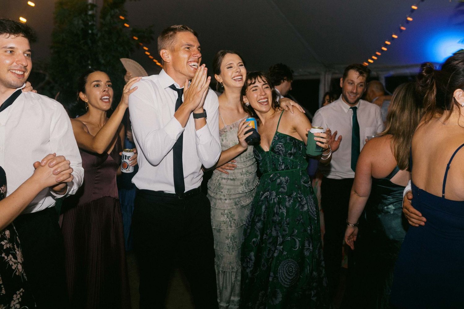 Wedding guests dance and celebrate on a dark dance floor illuminated by blue lighting at a reception party.
