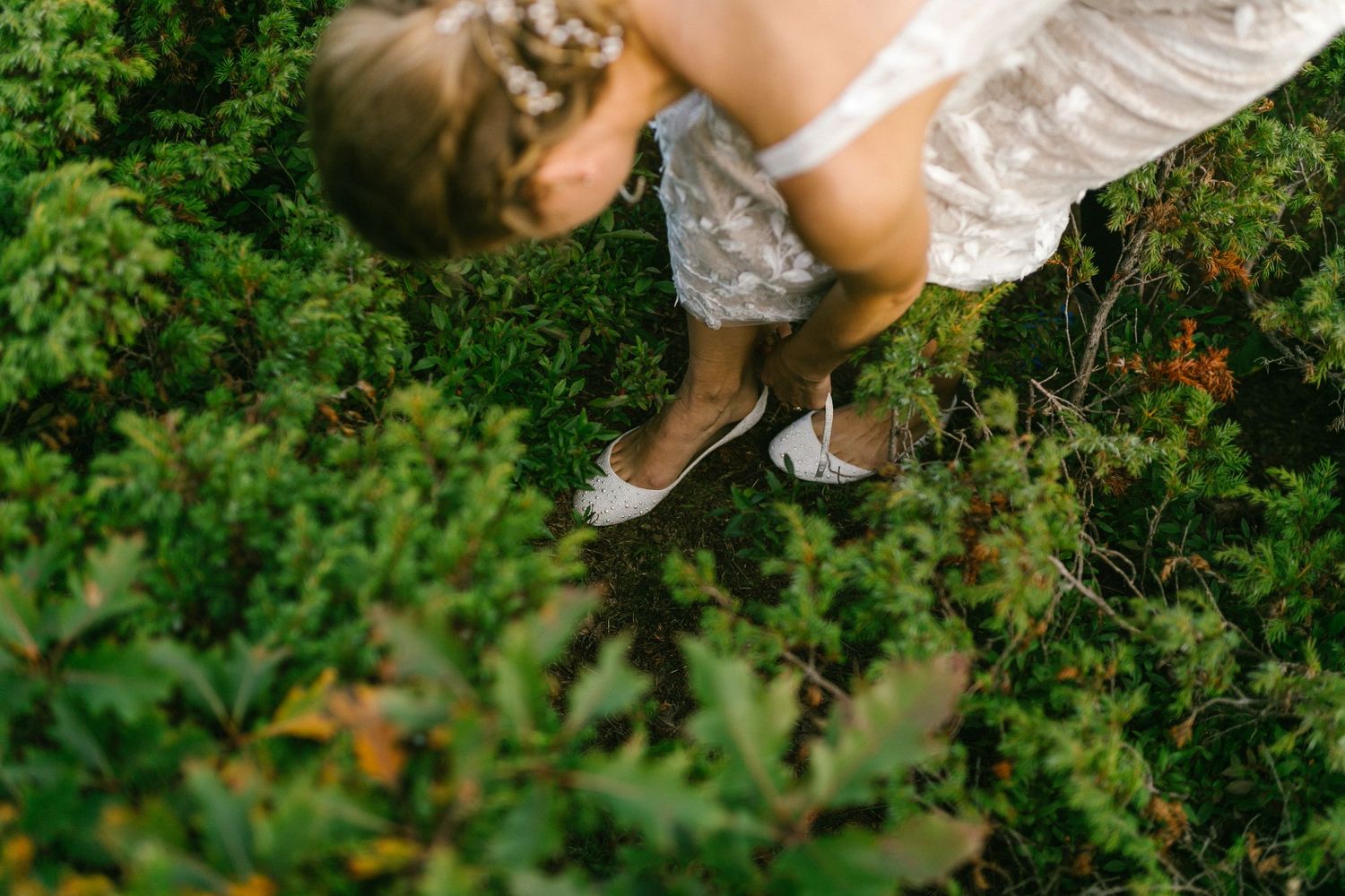 Looking down at vintage-inspired silver ballet flats and white lace dress amidst lush green garden foliage.