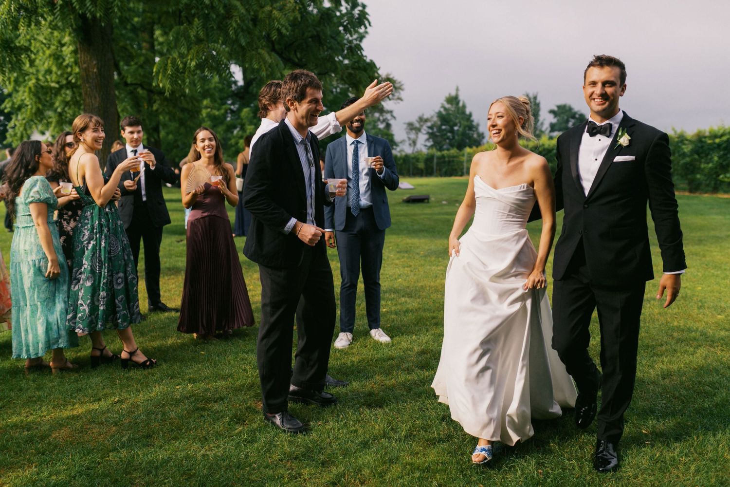 A wedding party celebrates on a lush green lawn with guests dancing and enjoying the outdoor festivities.