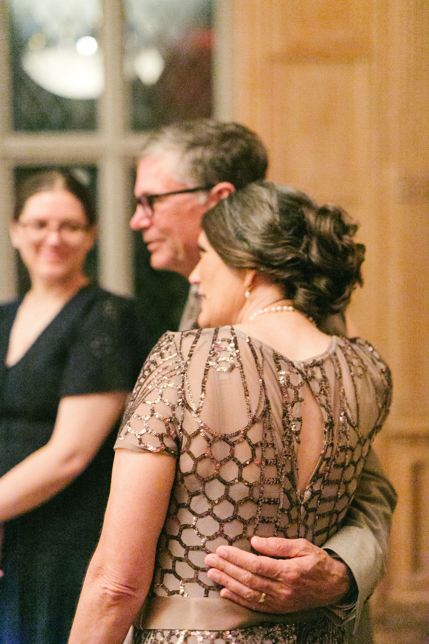 Elegant mother of the bride in beaded champagne gown embraces partner during wedding reception dance.