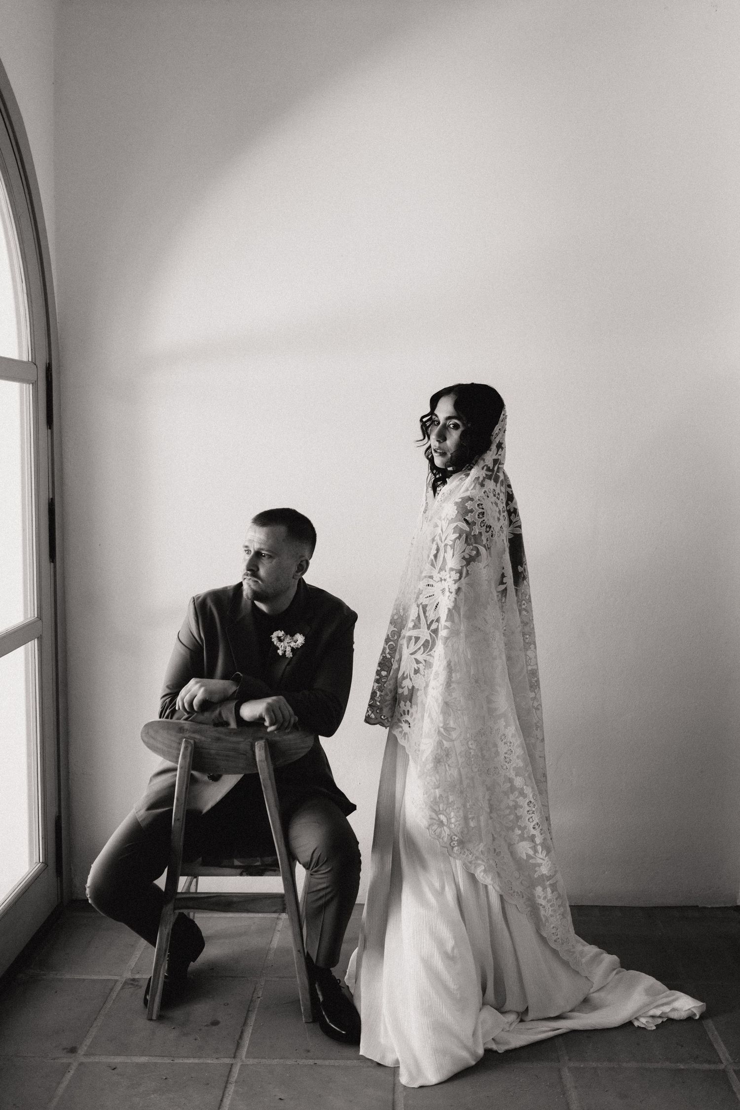 Black and white portrait of groom seated on stool beside standing bride in ornate lace cathedral veil.