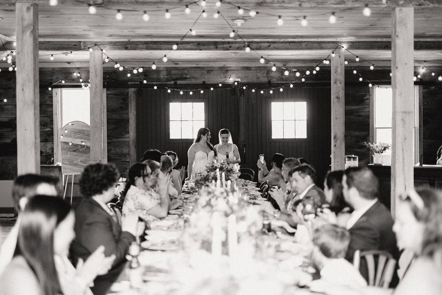 Black and white photo of wedding reception in rustic barn with string lights, long table, and couple standing at head.