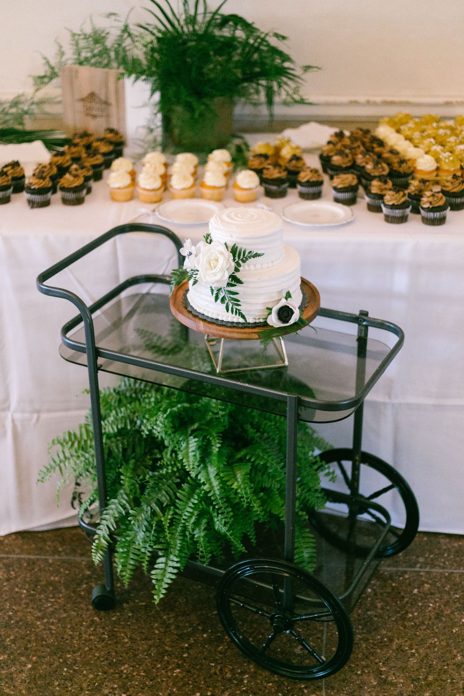 Vintage bar cart with ferns and dessert display featuring cupcakes and pastries on a white tablecloth.