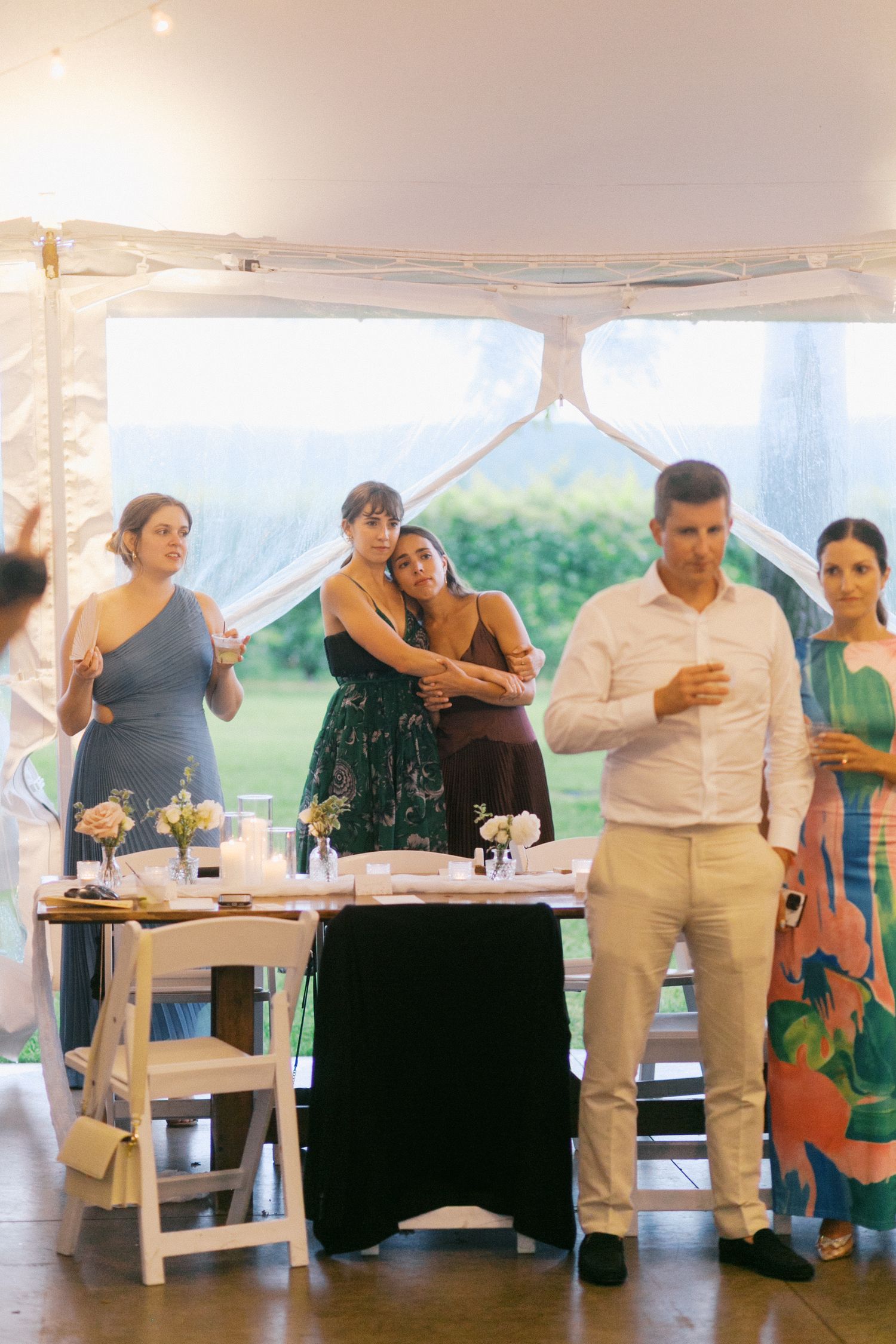 People gather under a white tent at an outdoor event with tables decorated with flowers and centerpieces.