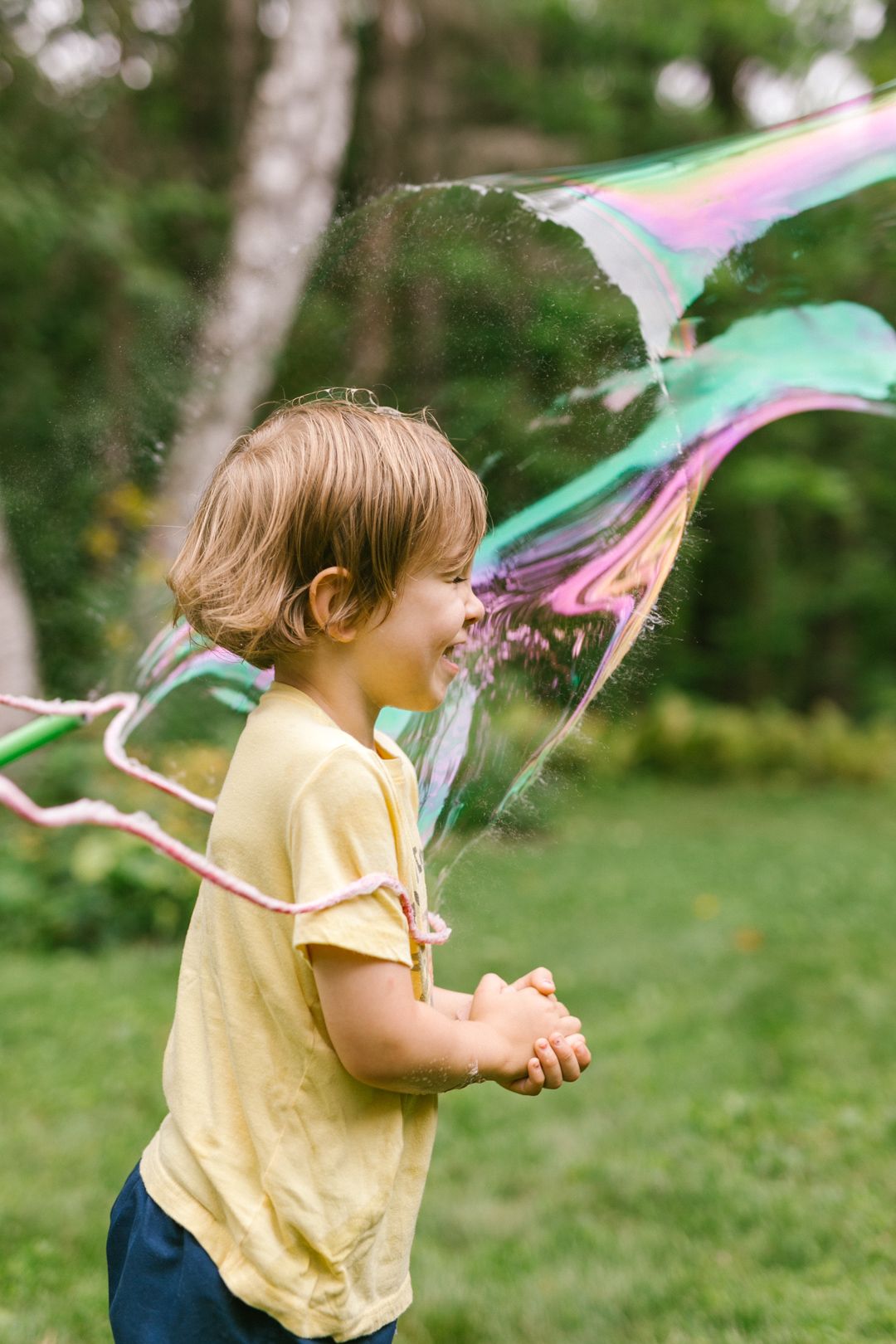 Happy toddler in yellow shirt laughing as a giant soap bubble bursts around them outdoors.