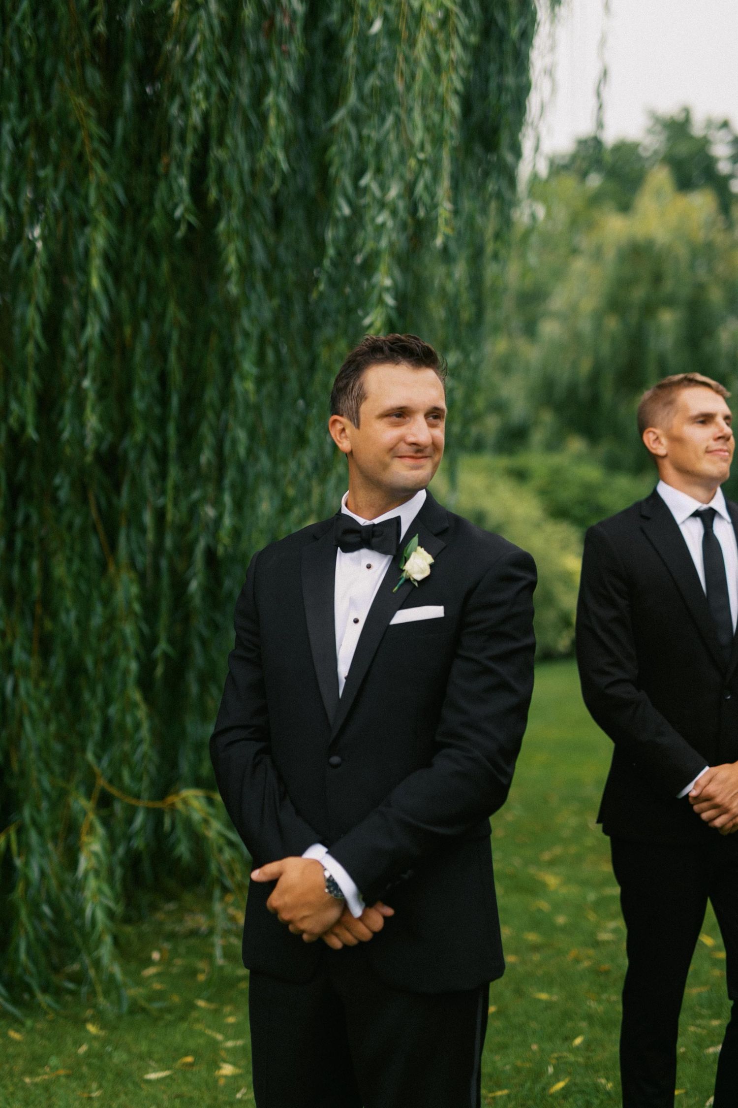 A groom in a black tuxedo smiles while watching his bride arrive under a willow tree during an outdoor wedding ceremony.