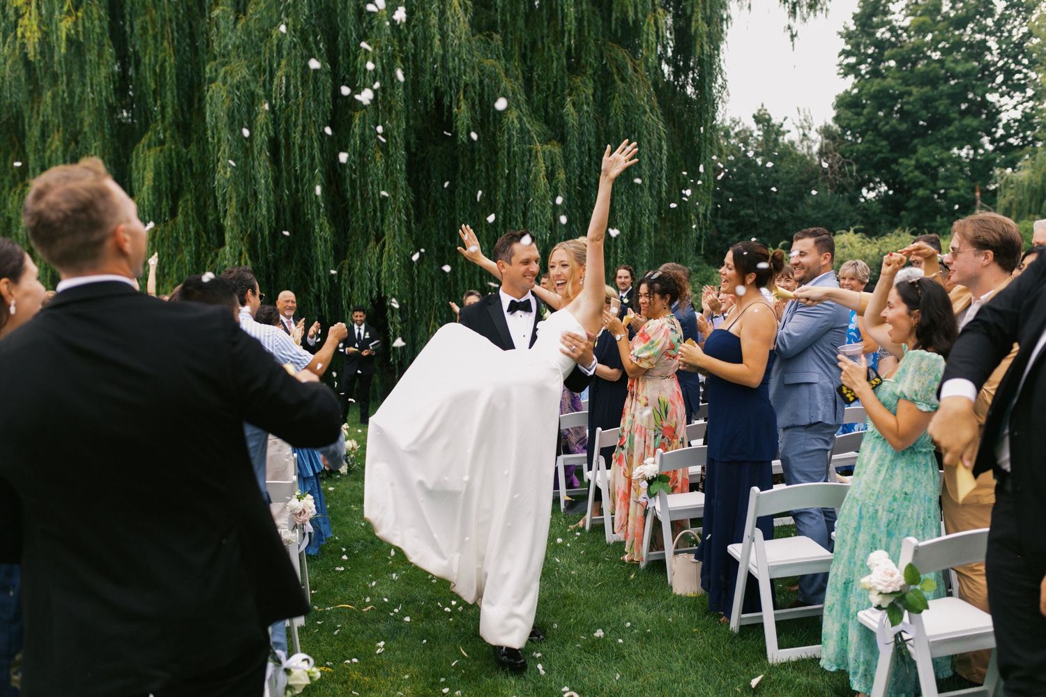 Guests toss flower pedals on a happy groom who carries his new bride back down the aisle from their outdoor ceremony under a willow tree.