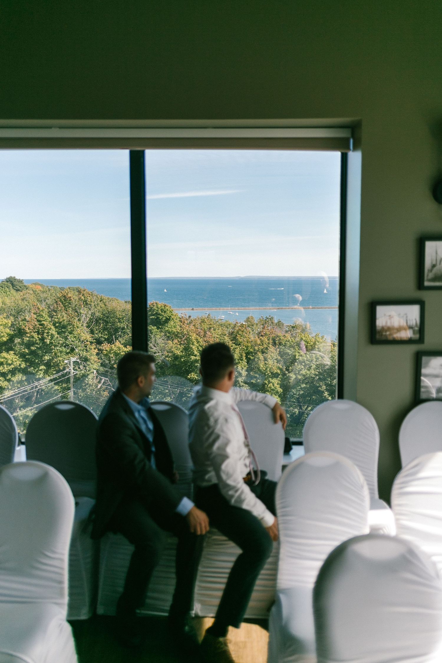 Wedding venue interior with white chair covers and large windows overlooking an ocean view on a sunny day.