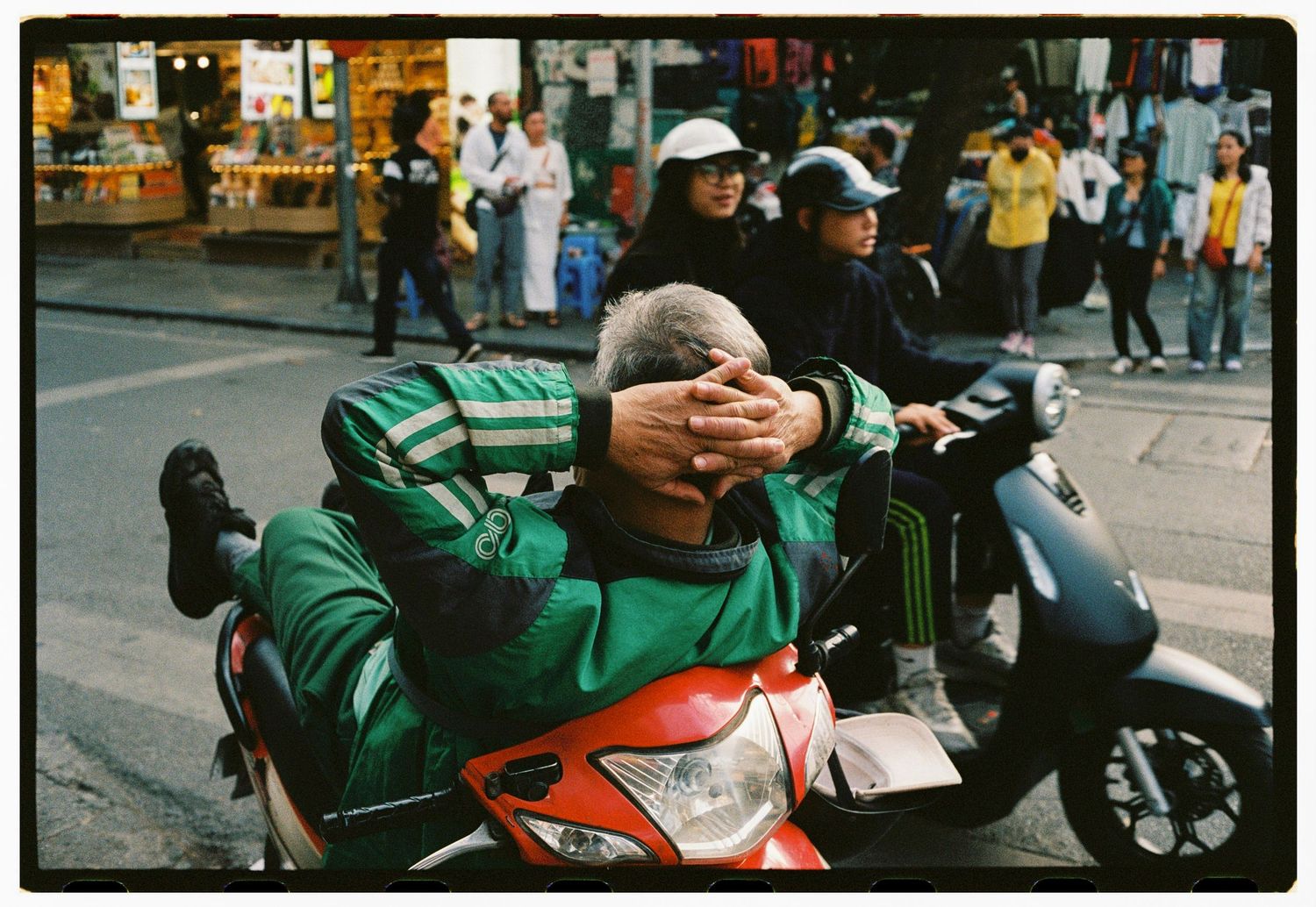 Man relaxing on a red motorcycle in a busy street, with other riders and pedestrians in the background.