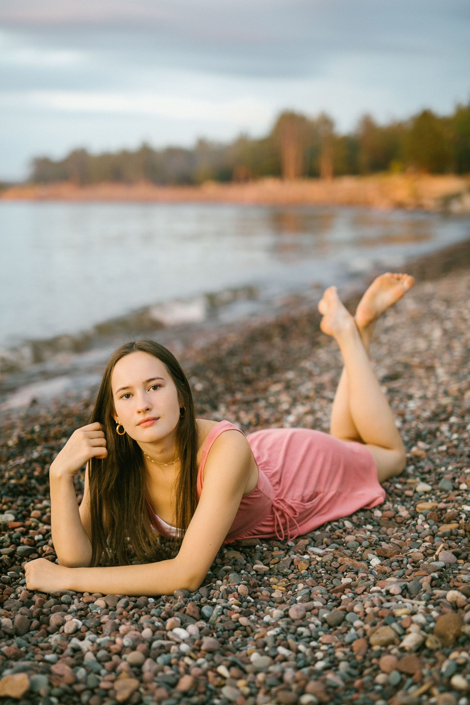 Young brunette woman in pink dress lying on rocky lakeside beach at sunset.