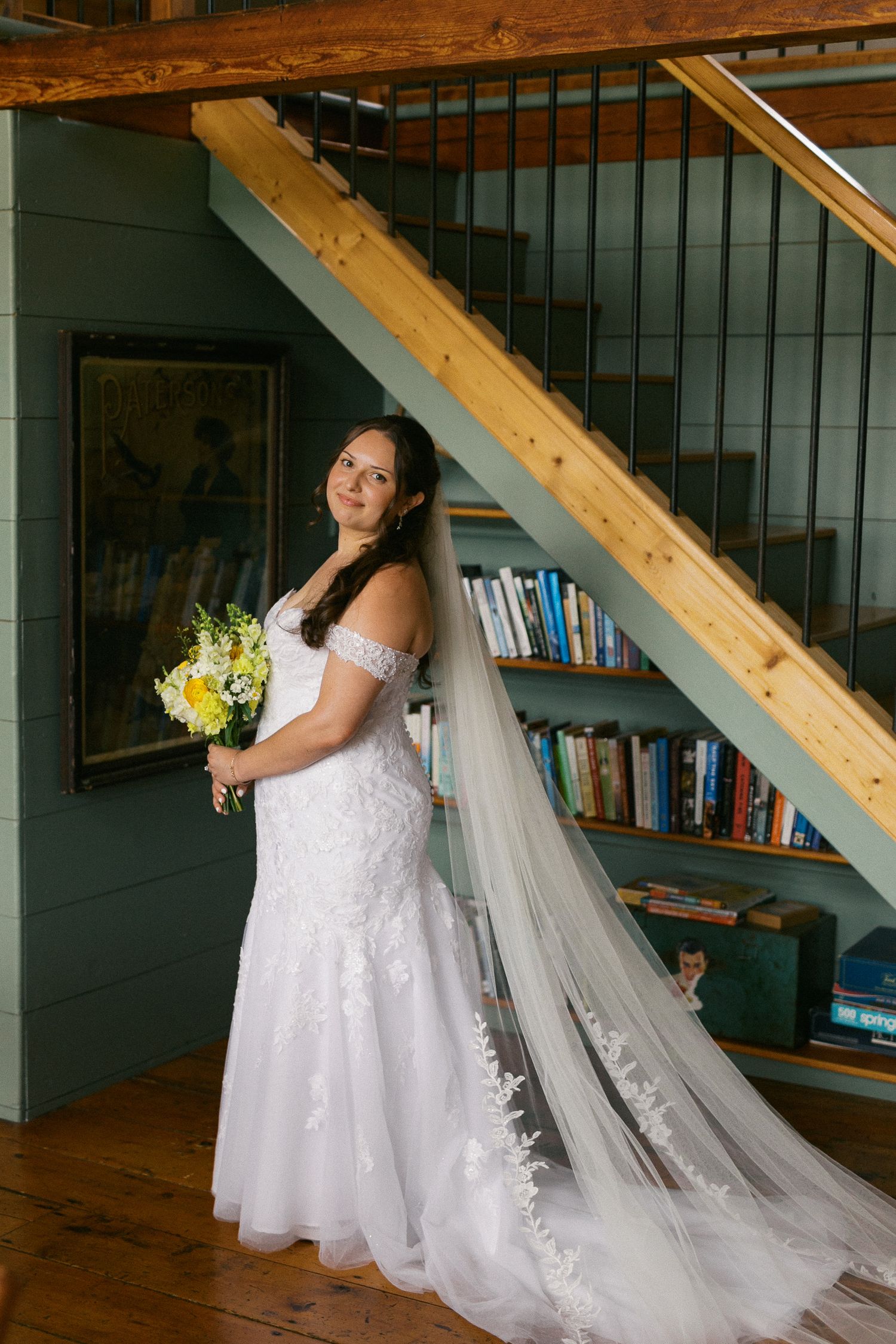 Bride in off-shoulder lace mermaid gown with cathedral veil, holding yellow bouquet beside rustic wooden staircase.