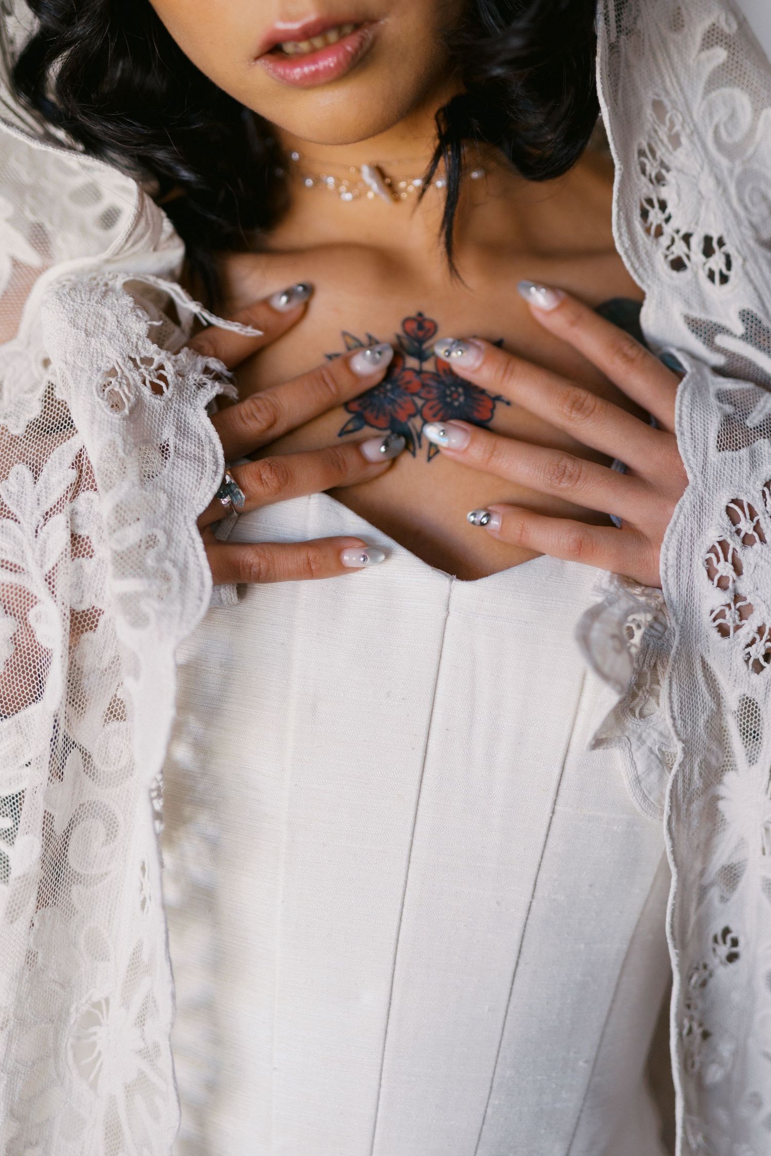 Bride in white corset and lace robe with floral chest tattoo and decorated nails poses elegantly.