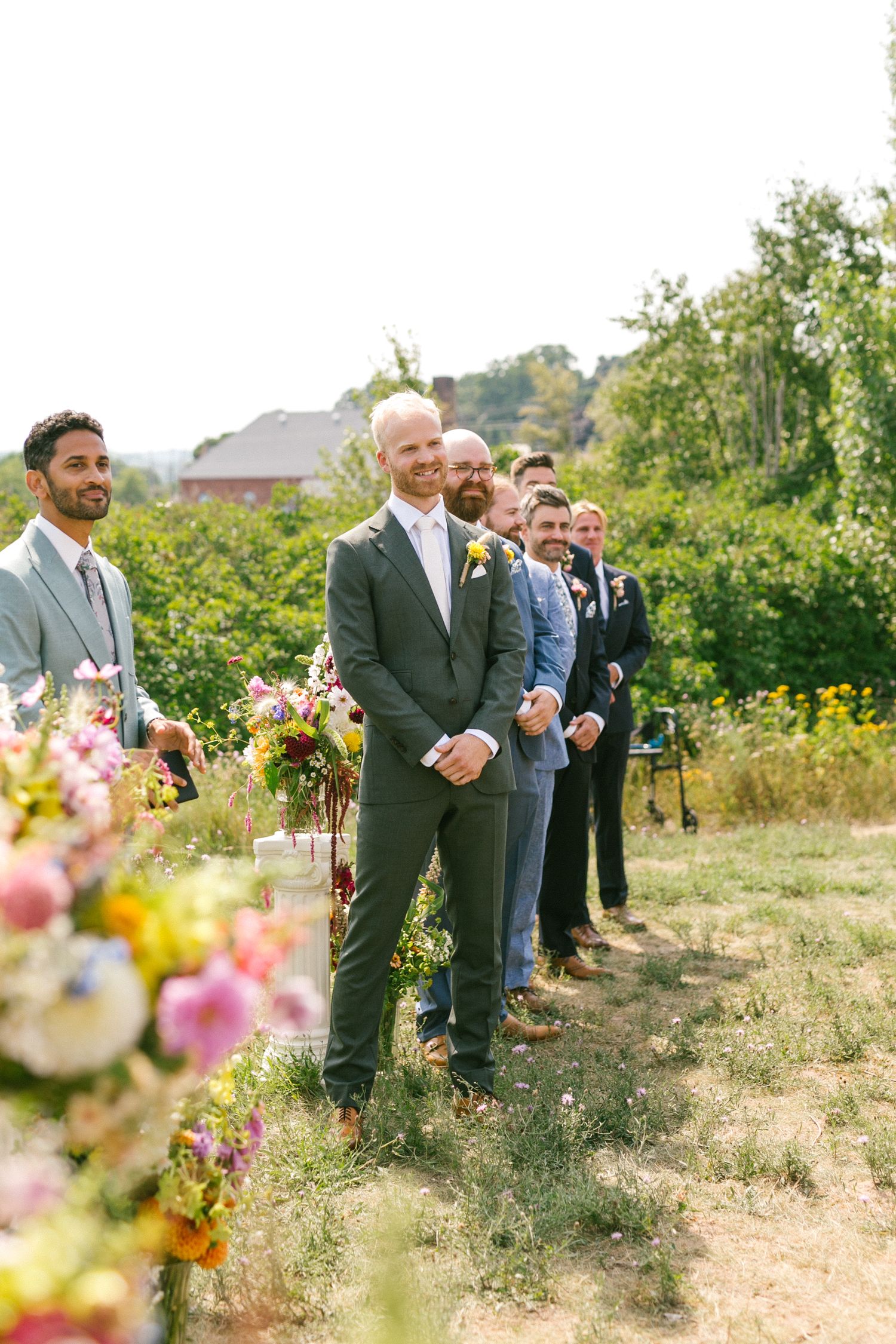 Groomsmen stand in line at an outdoor wedding ceremony with wildflowers in the background.