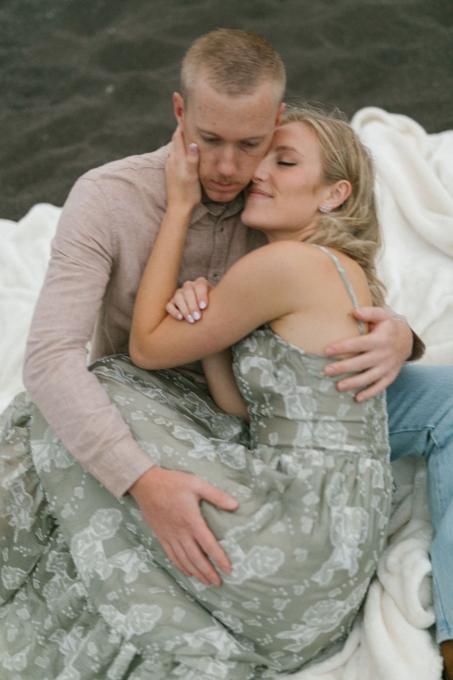 Intimate couple cuddles on a white blanket outdoors, sharing a tender moment together.