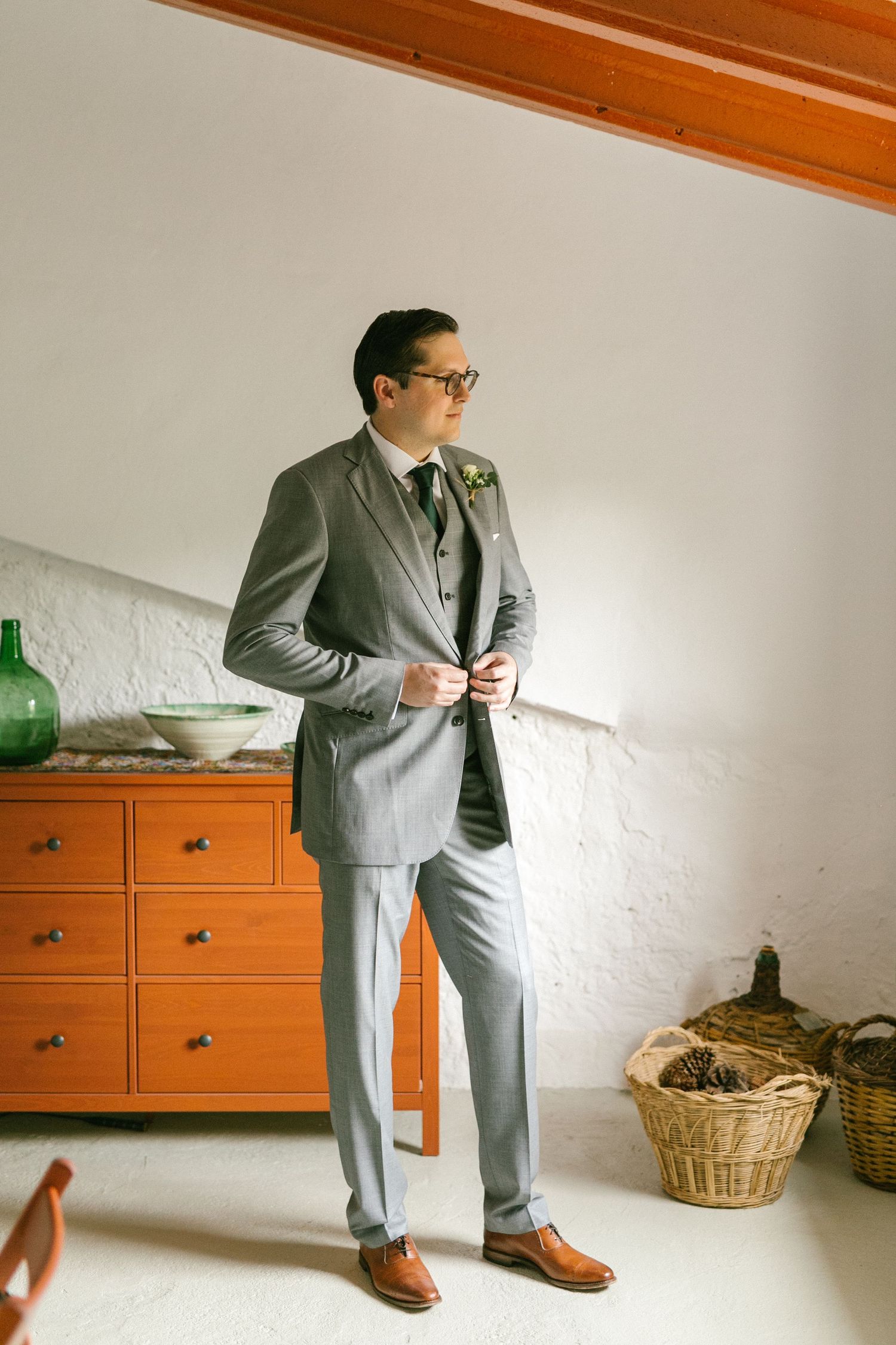 A spanish groom wearing a light gray three-piece suit with brown dress shoes stands near an orange dresser in a bright interior space.