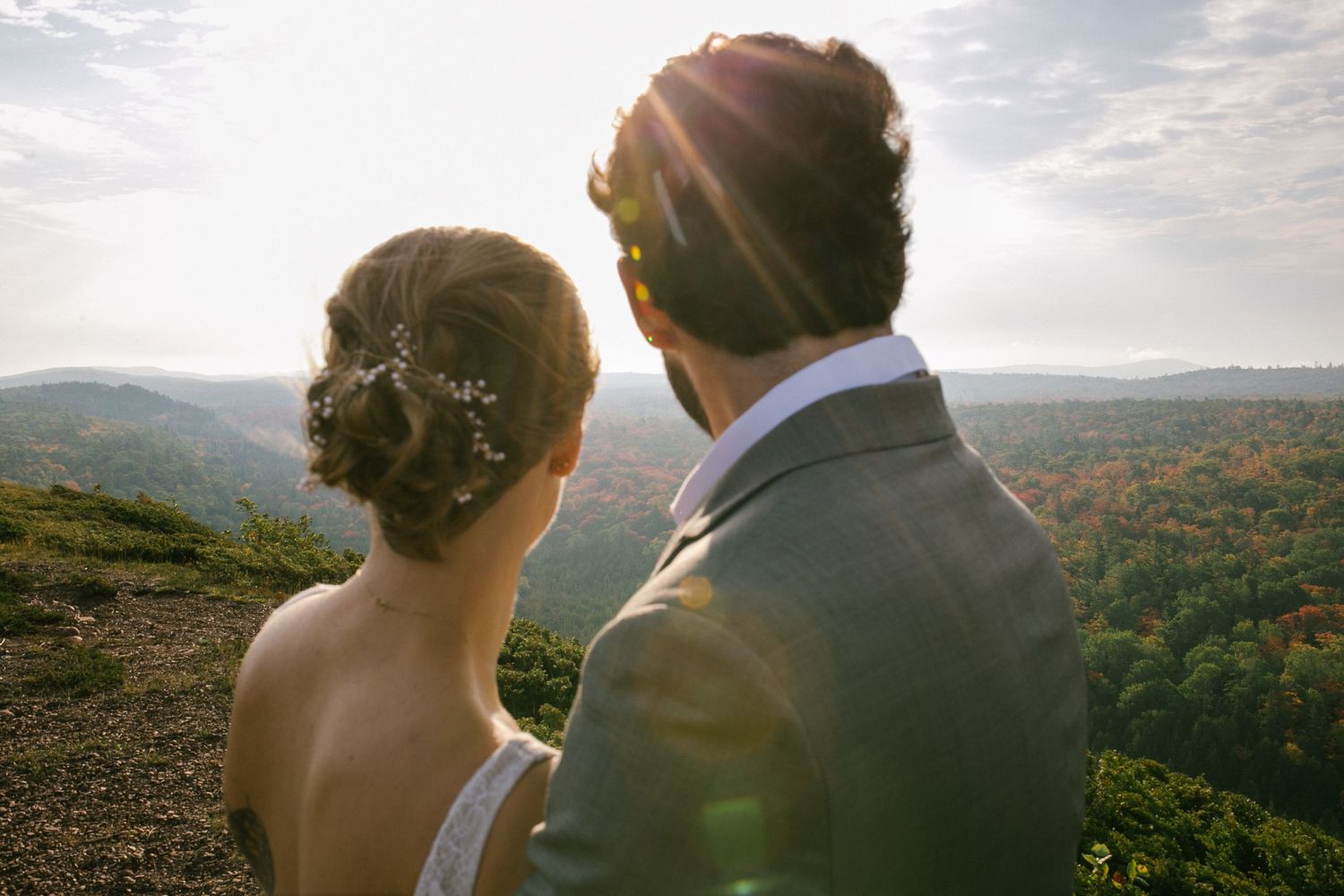 A couple in wedding attire gazes at a scenic mountain vista during golden hour, with sunlight streaming through the trees.