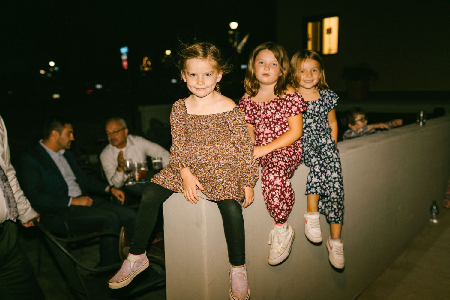 Young friends in patterned dresses sit on a ledge during an evening celebration.