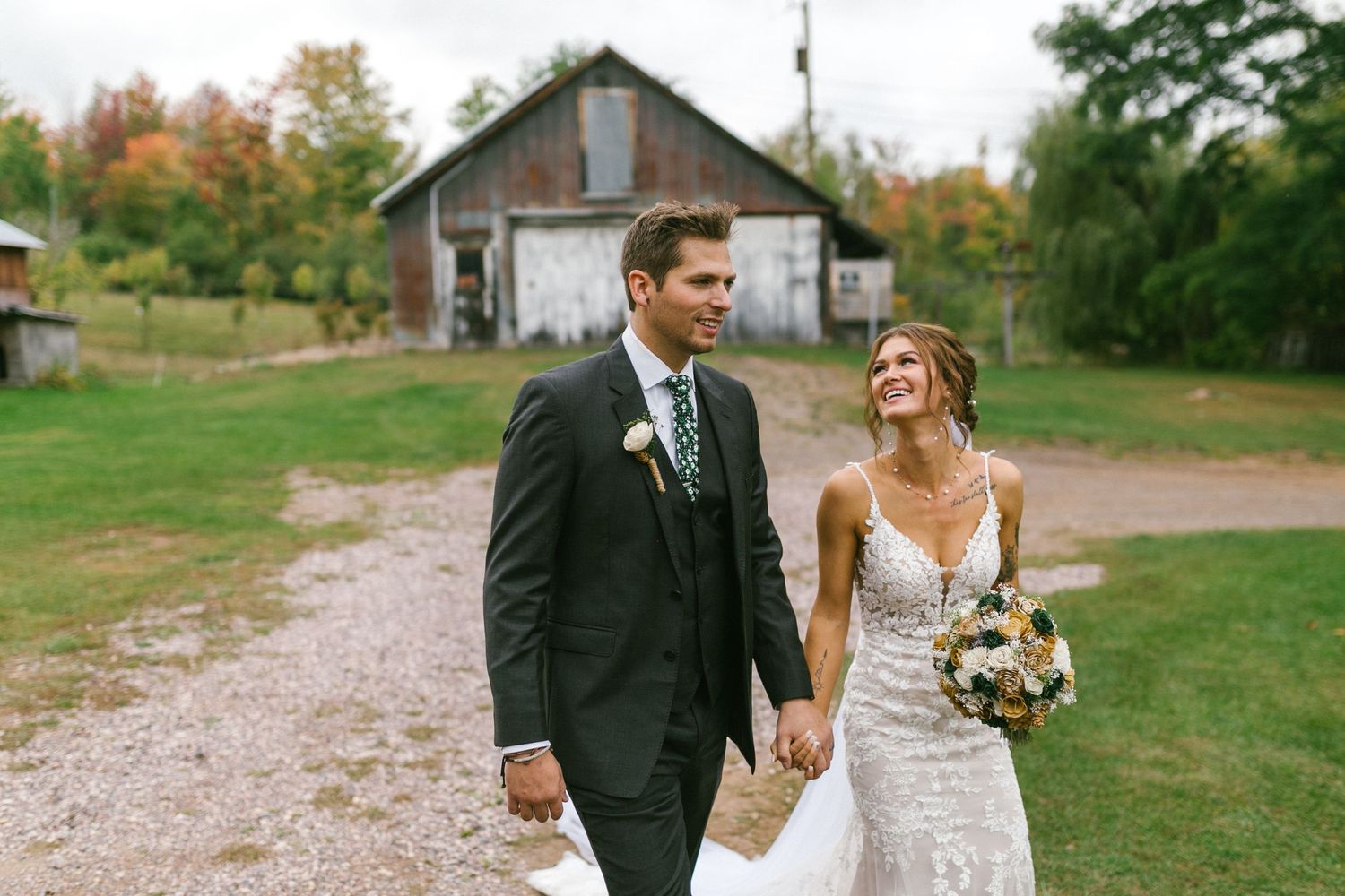 A couple in wedding attire walks hand in hand along a gravel path with a rustic barn in the background.