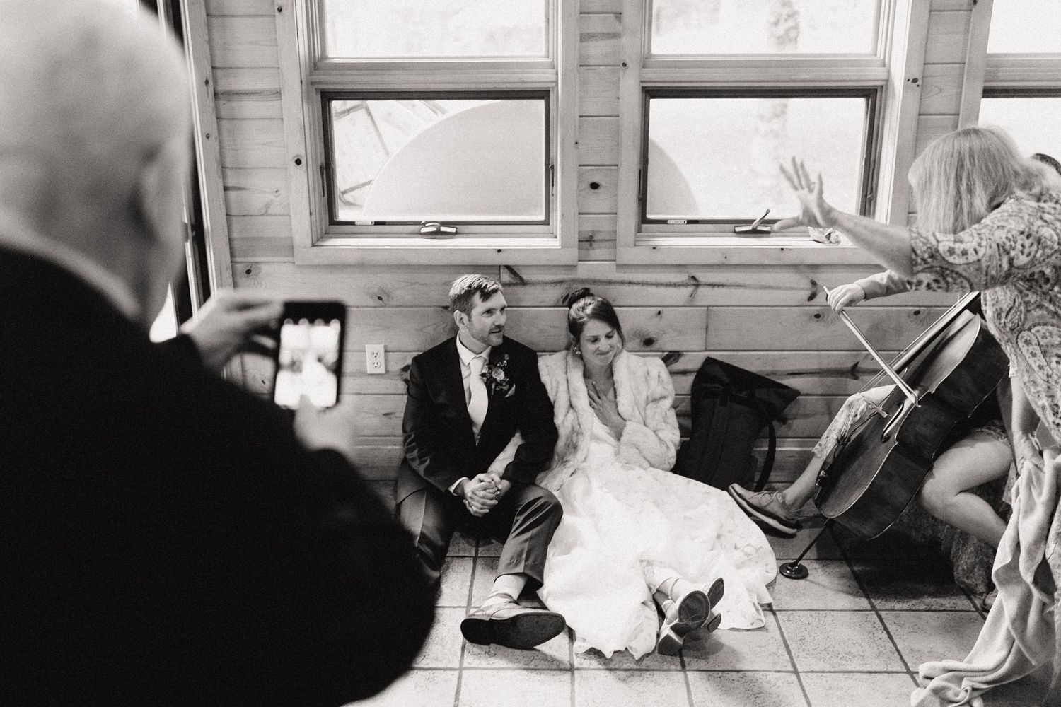 A candid black and white wedding photo showing a couple seated on a window ledge while someone takes their picture.