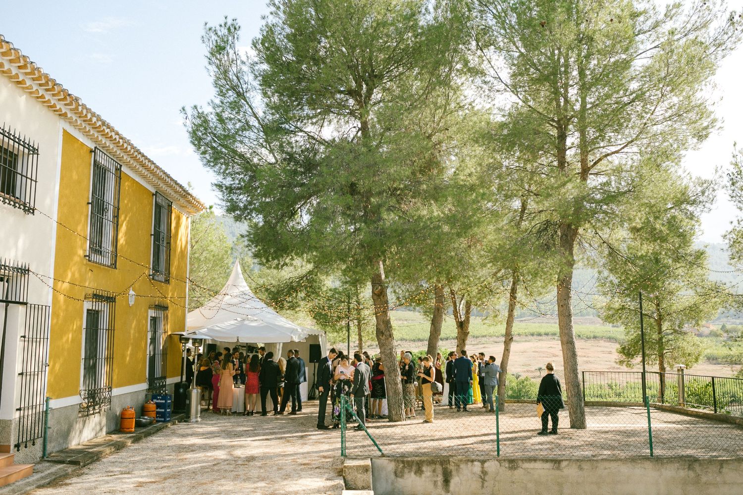 Guests gather outside a yellow Mediterranean building with tall pine trees during an outdoor event.