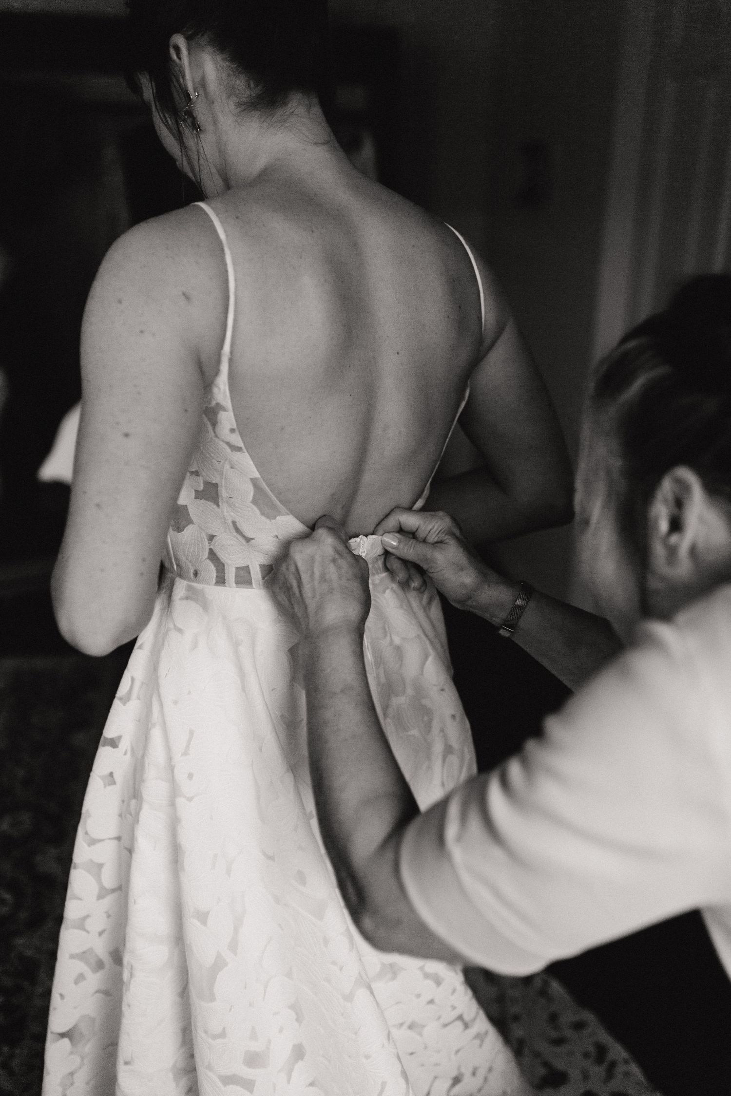 Black and white photo showing someone helping to fasten buttons on the back of a white lace dress.