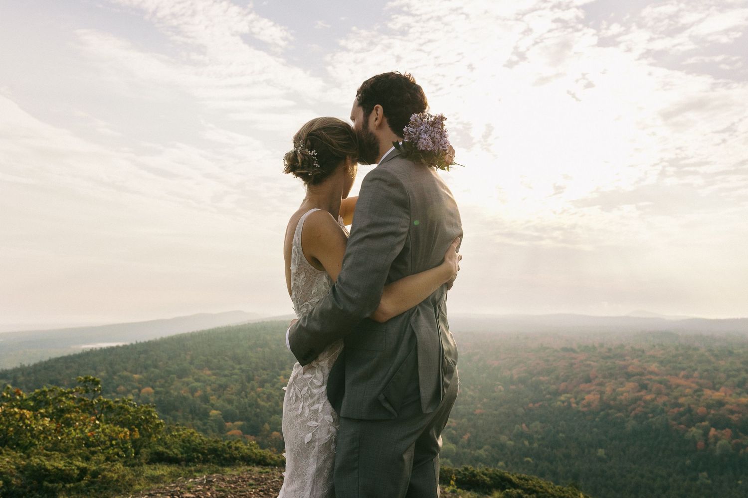 Bride and groom embrace while overlooking stunning forested valley landscape at sunset.