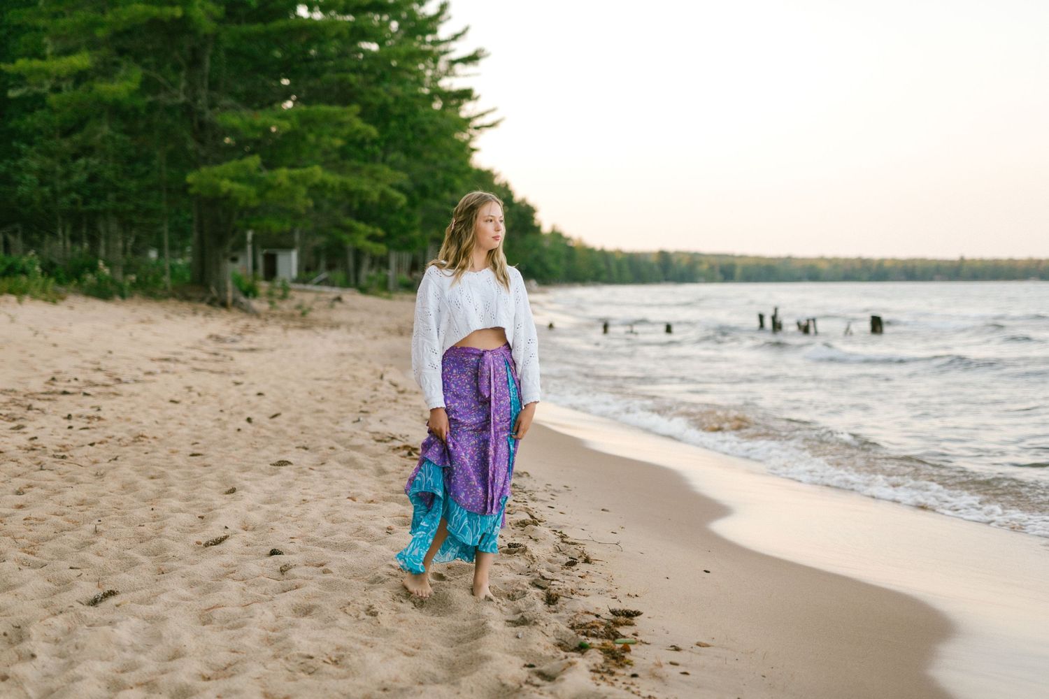 Woman in white top and purple pants walking barefoot on sandy beach.