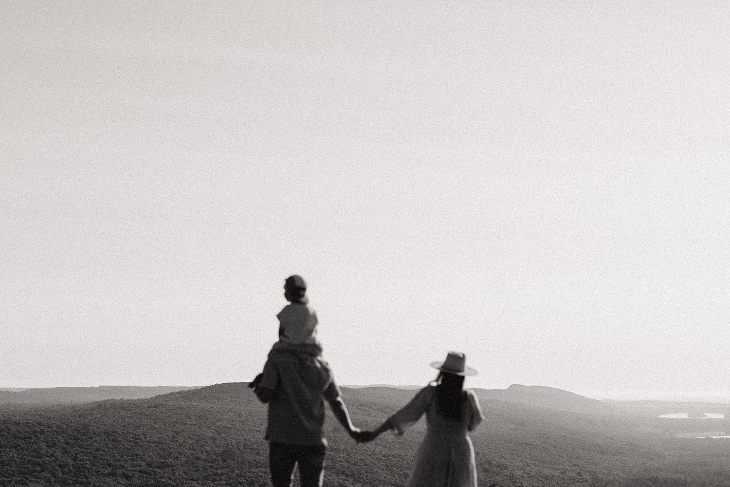 Black and white photo of a family of three walking on a hilltop, father carrying child on shoulders.