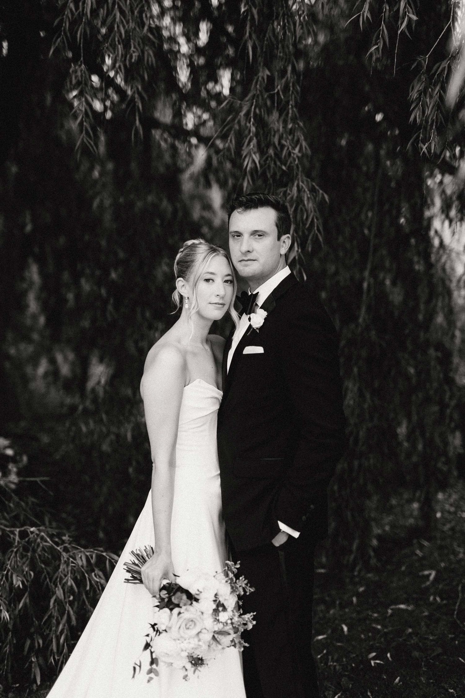 A black and white wedding portrait under willow trees with a couple in formal attire holding a floral bouquet.