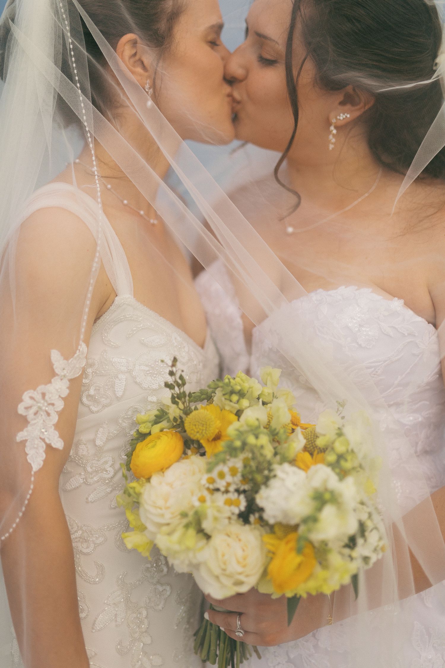 Two brides in white lace gowns kissing under a veil, holding a yellow and white floral bouquet.