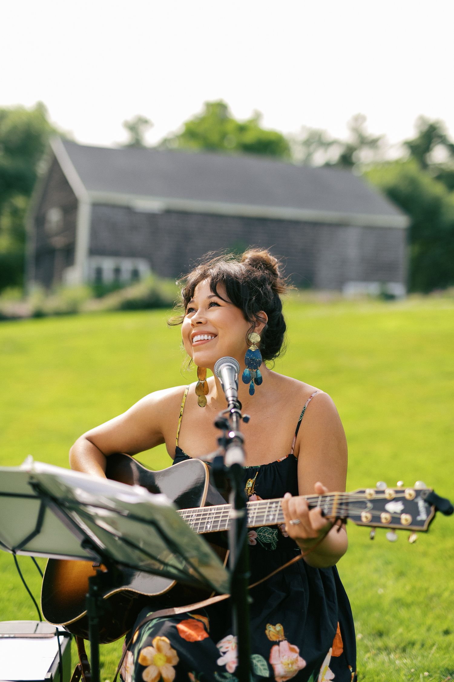 Smiling woman playing acoustic guitar outdoors at a sunny garden event, wearing a floral dress and colorful earrings.