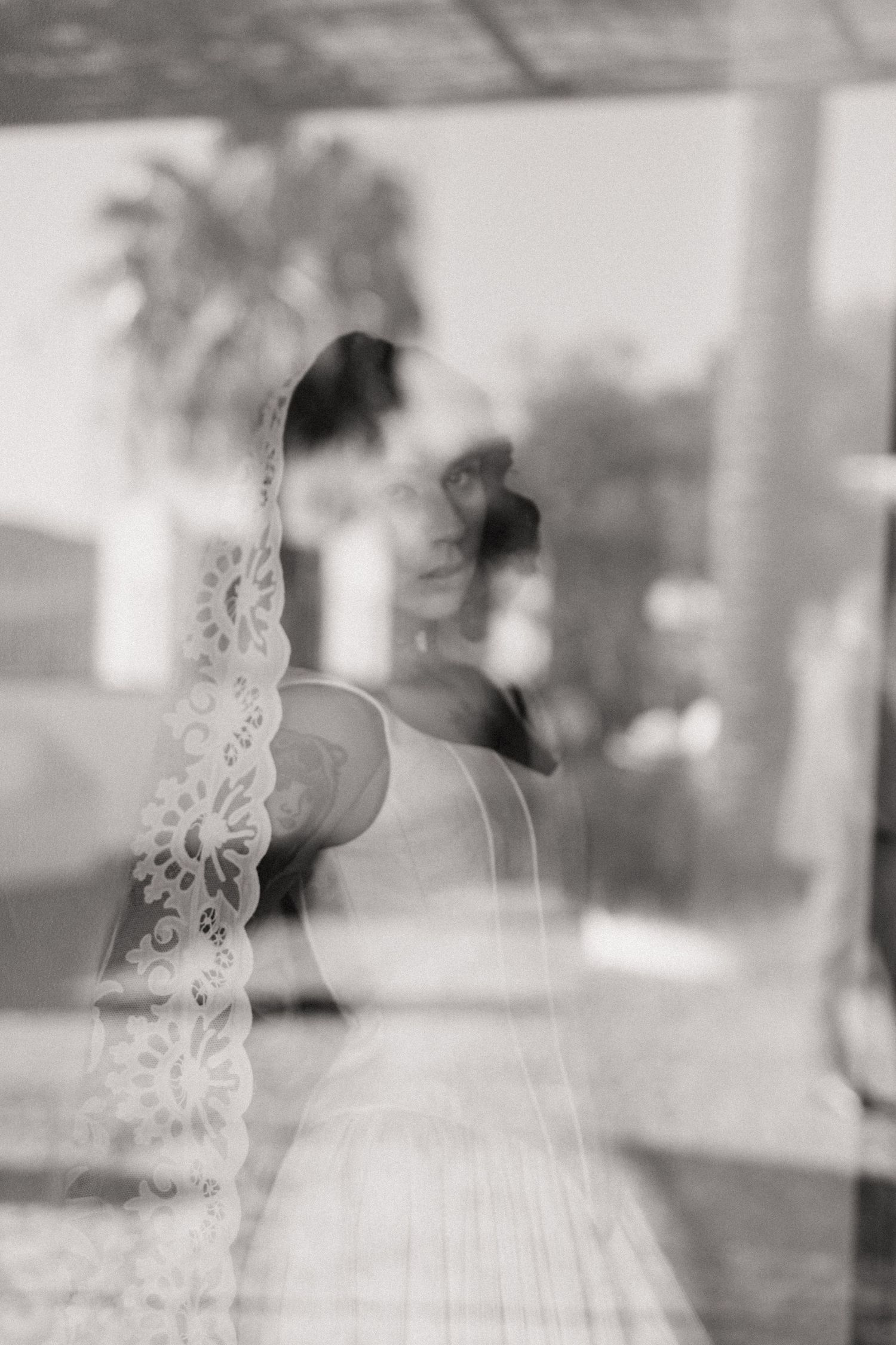 Artistic black and white reflection photo of bride in lace veil through glass window with palm trees.