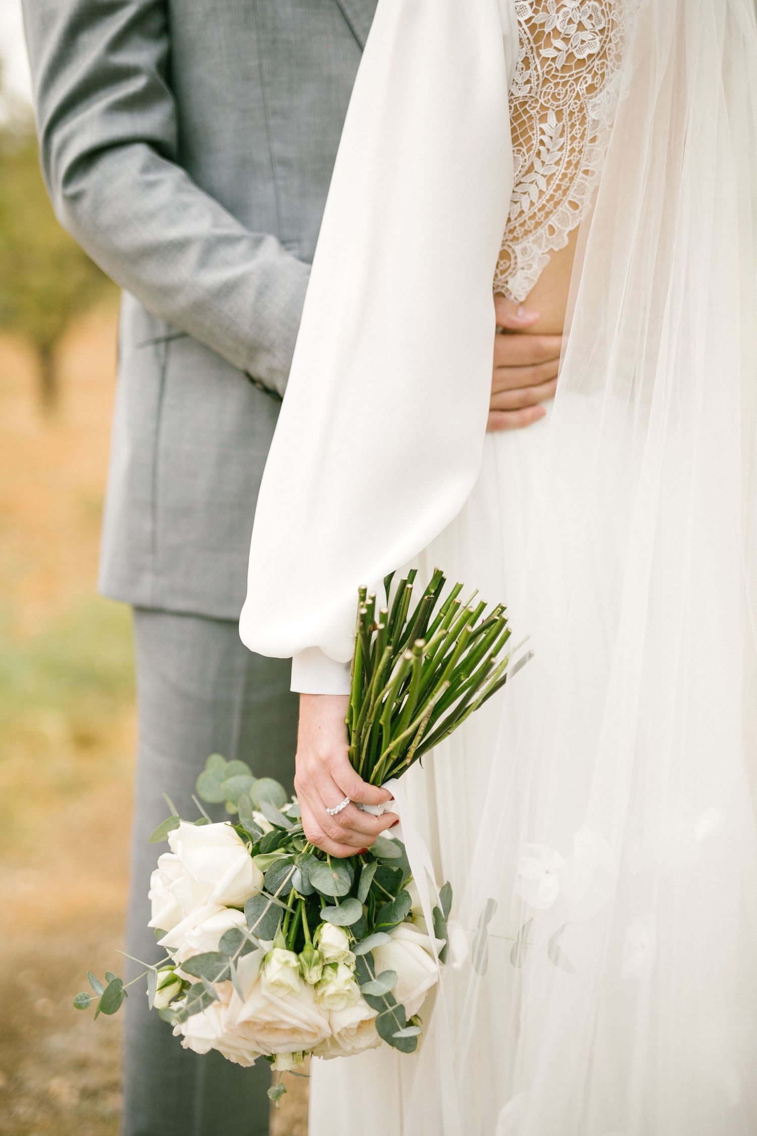 A bridal bouquet of white roses and greenery is held against a flowing white wedding dress and gray suit.