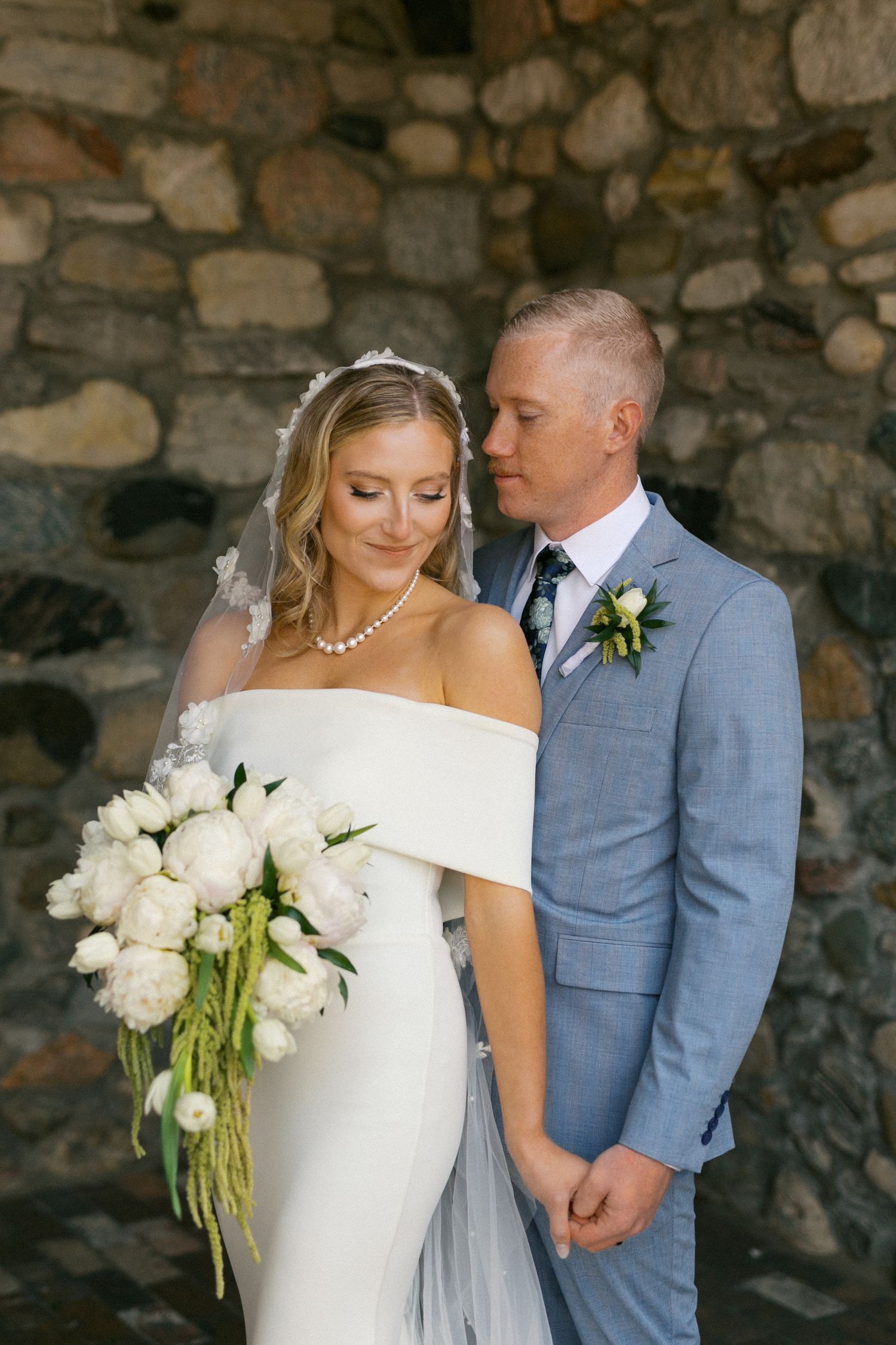 Elegant bride in off-shoulder white dress holding a lush bouquet of white flowers against a stone wall.