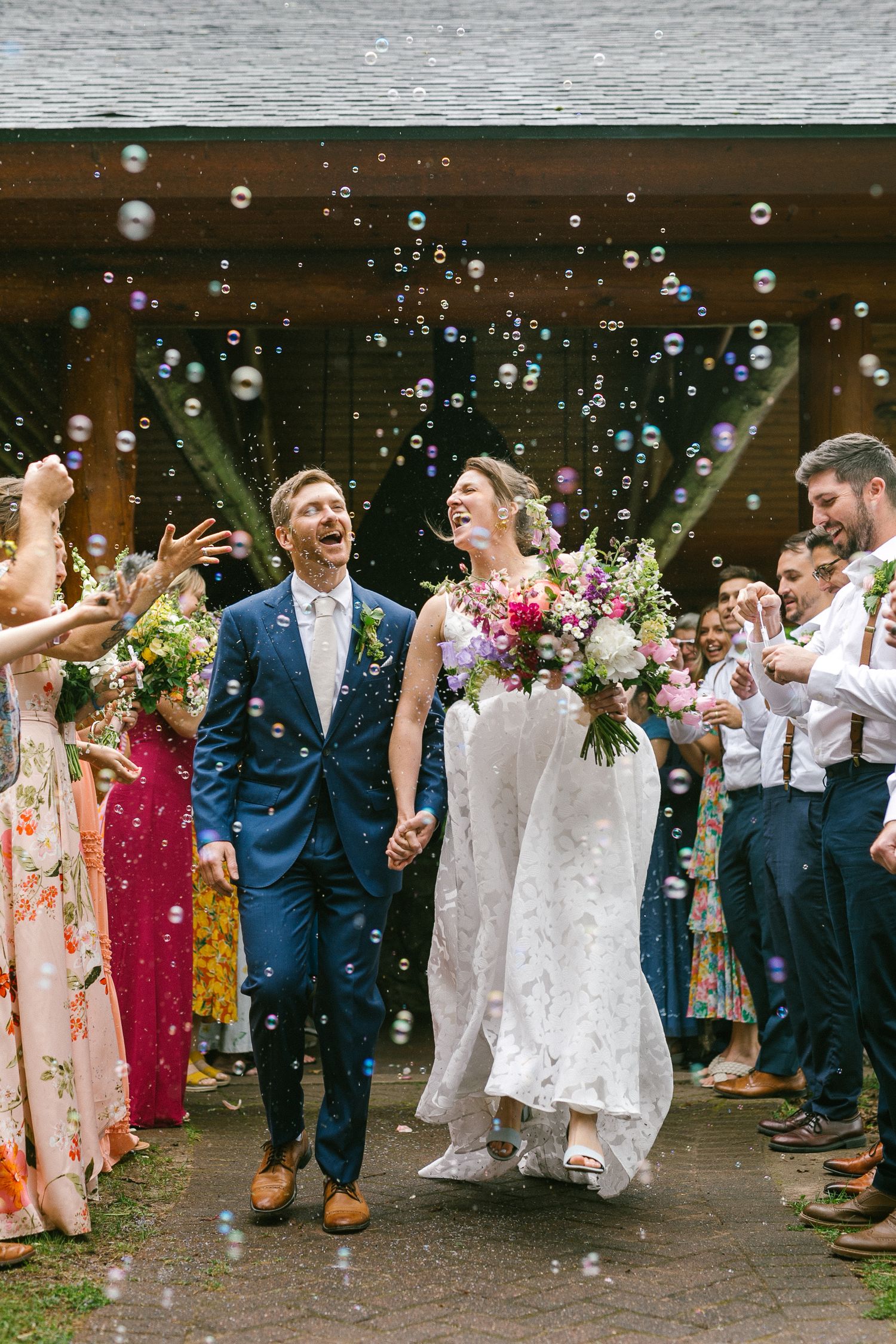 Wedding guests blow bubbles as couple celebrates during outdoor ceremony, surrounded by floral decorations and festivities.