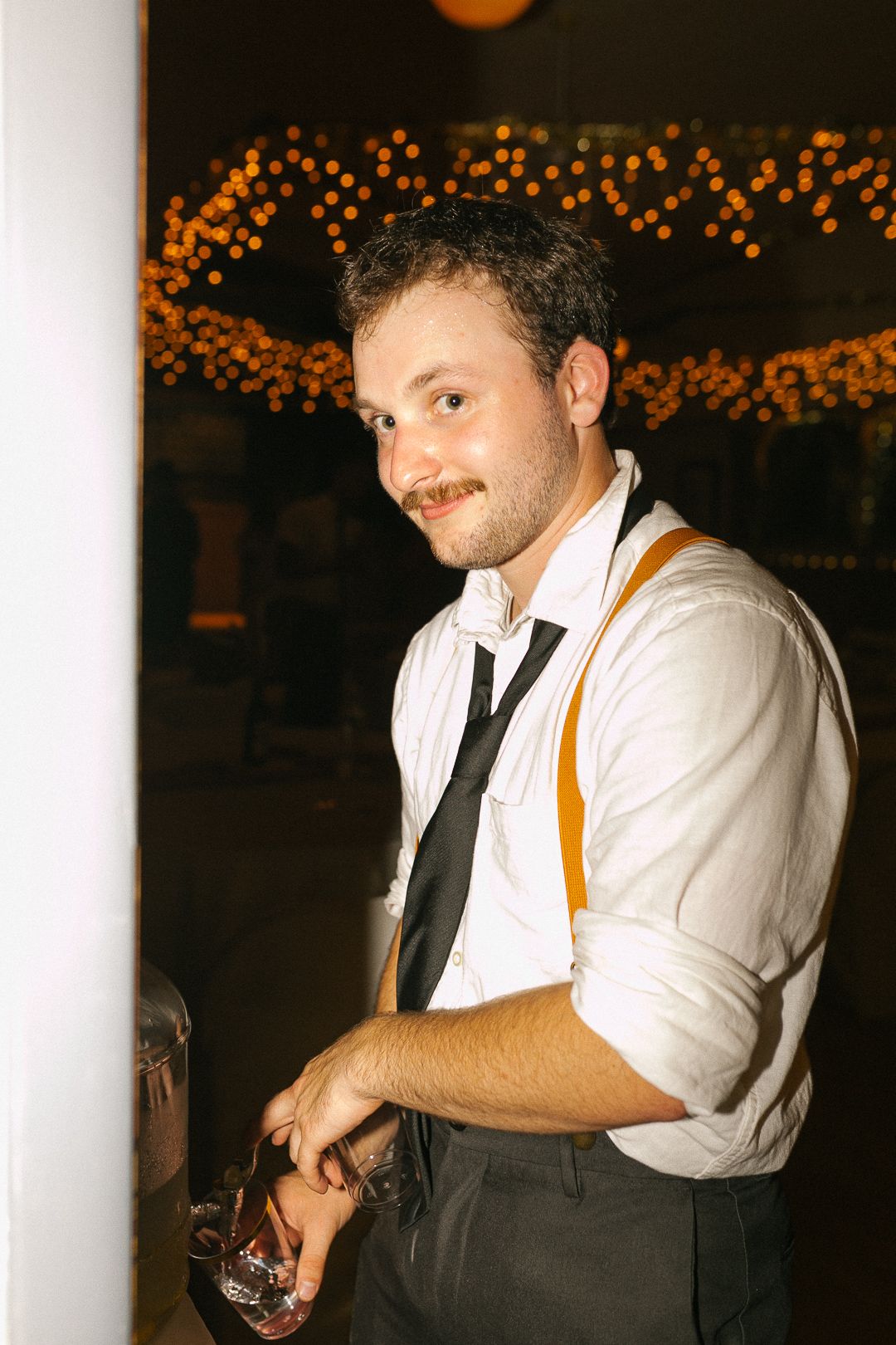 A bartender in a white shirt and suspenders serves drinks at an evening reception with twinkle lights in the background.