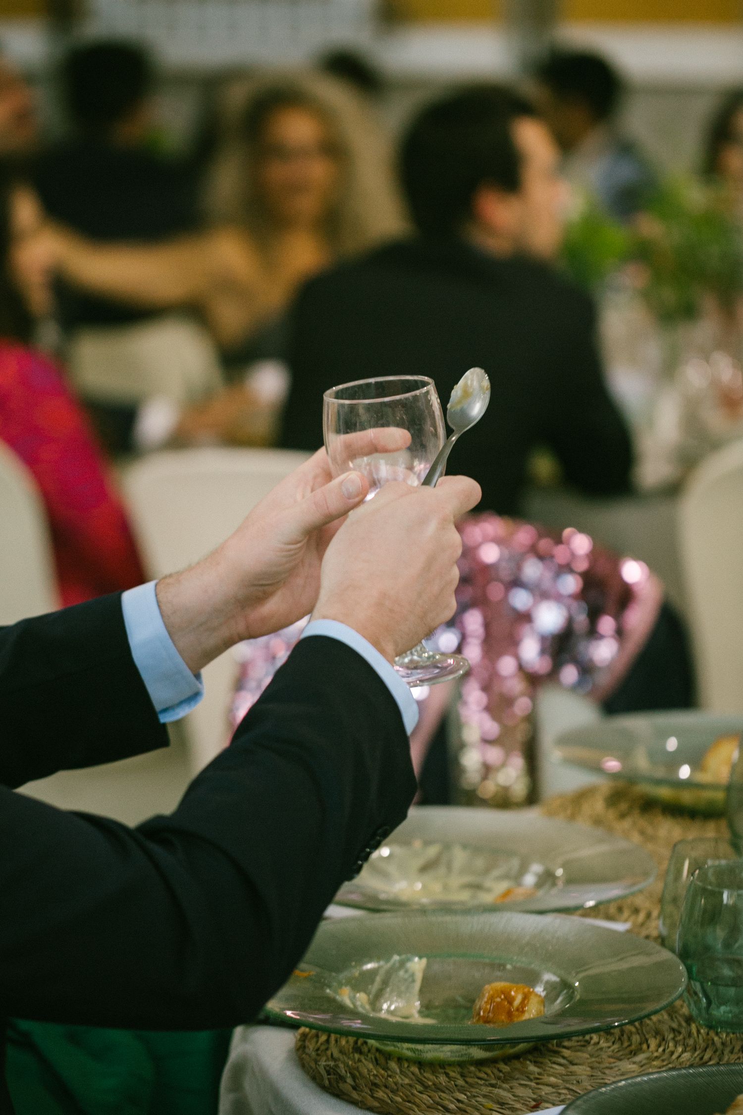 A guest holds up a wine glass at a formal dining event with table settings and other guests in the background.