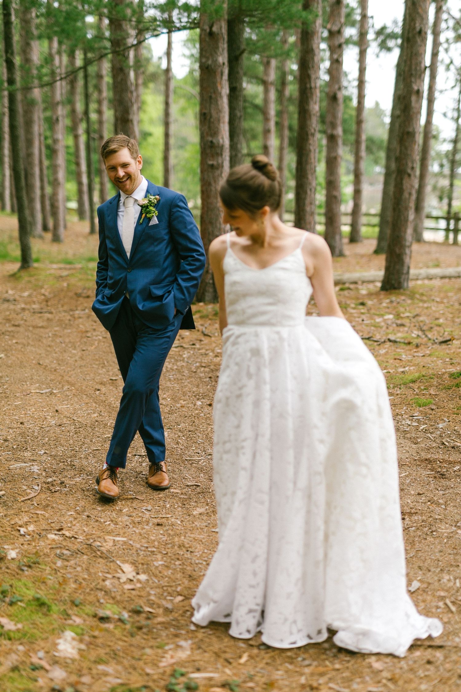 A couple in wedding attire walking through a pine forest with the bride in white and groom in navy blue.
